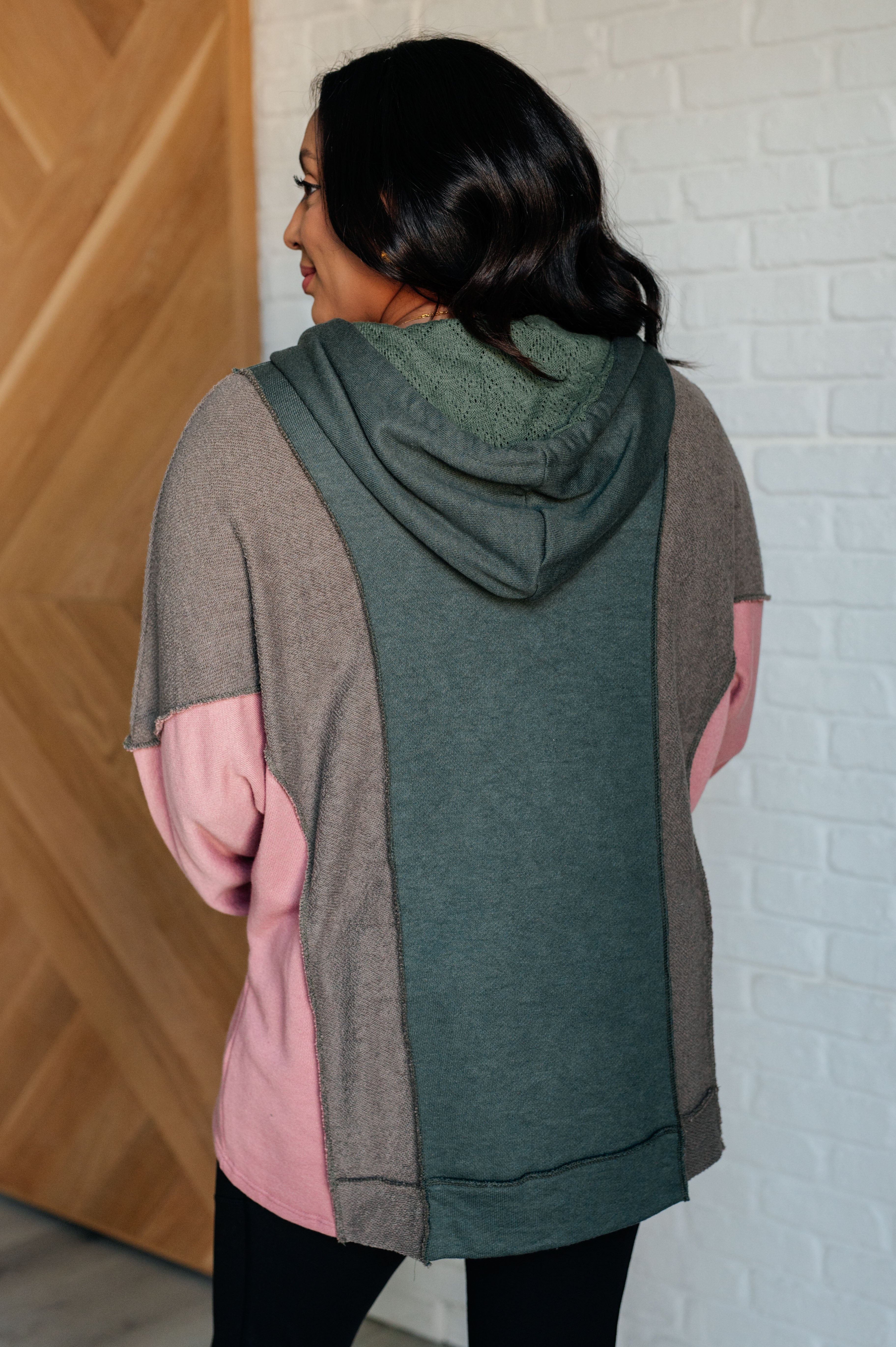 Woman wearing a relaxed fit hoodie with sage green, muted brown, and soft pink color blocks, shown from the back indoors near a wooden door and white brick wall.