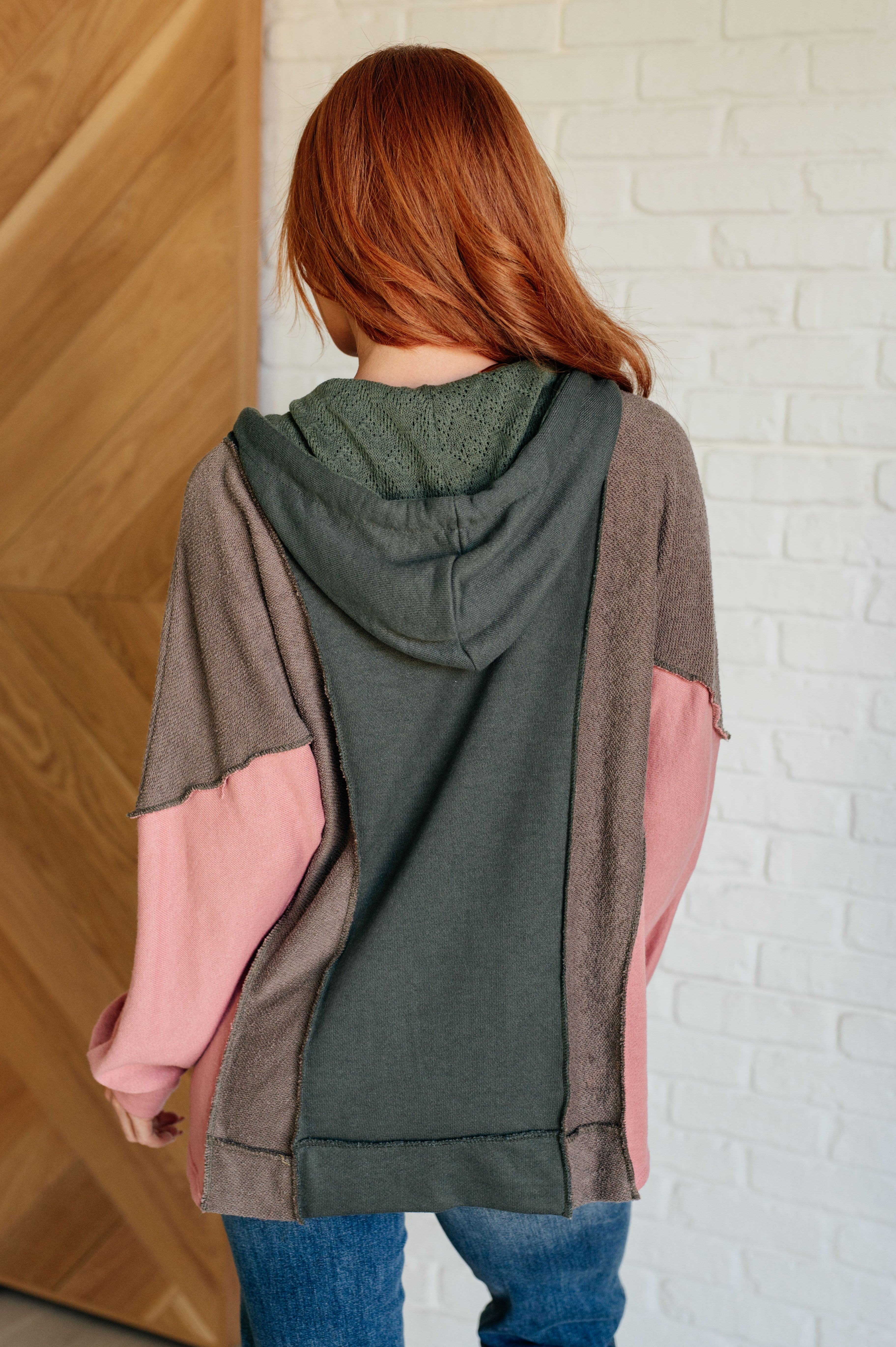 Rear view of a person with long red hair wearing a relaxed fit color block hoodie in sage green, muted brown, and soft pink standing indoors by a white brick wall and wooden door.