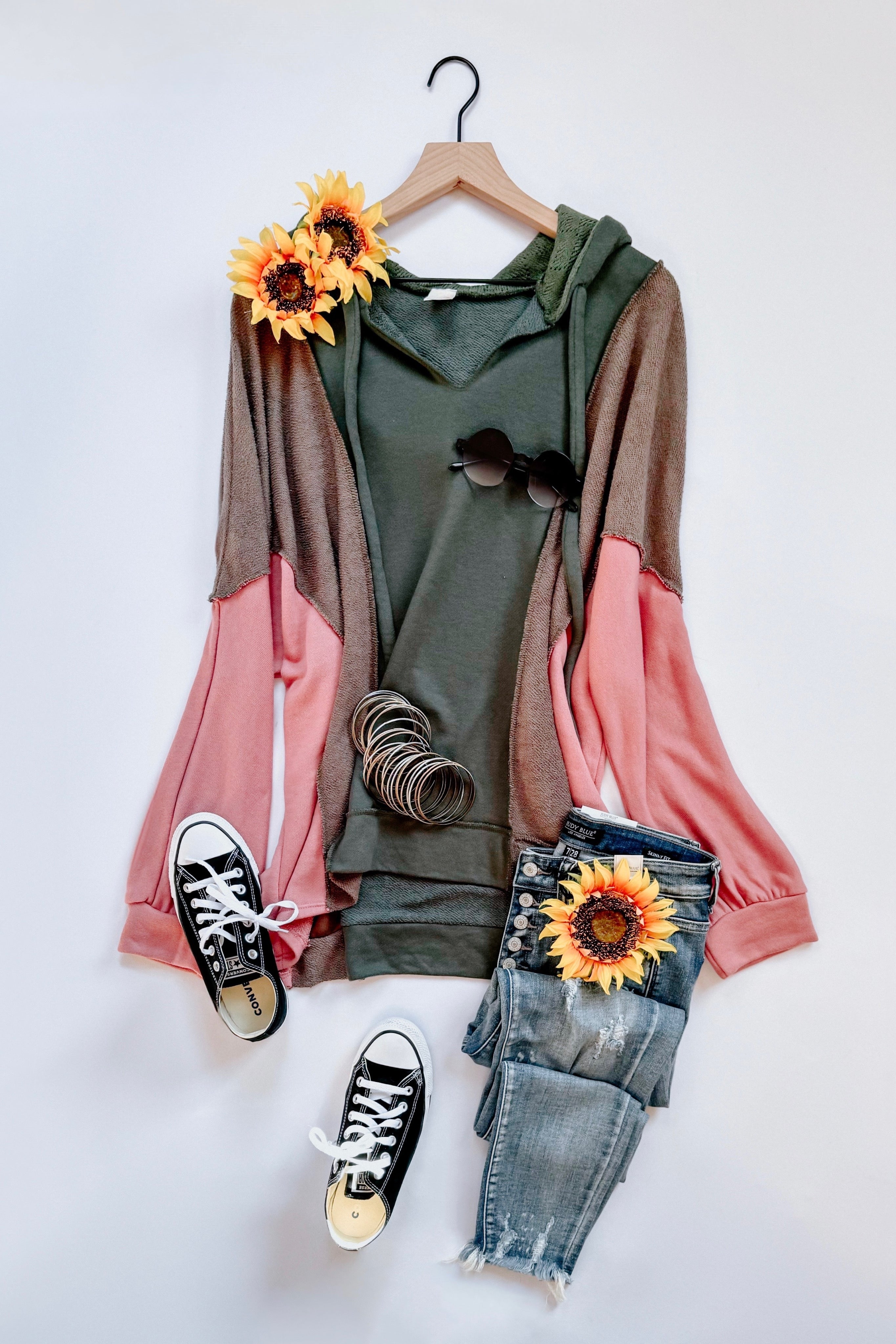 Relaxed fit hoodie in sage green with muted brown and soft pink color blocking, styled with distressed blue jeans, black sneakers, and sunflower accents.