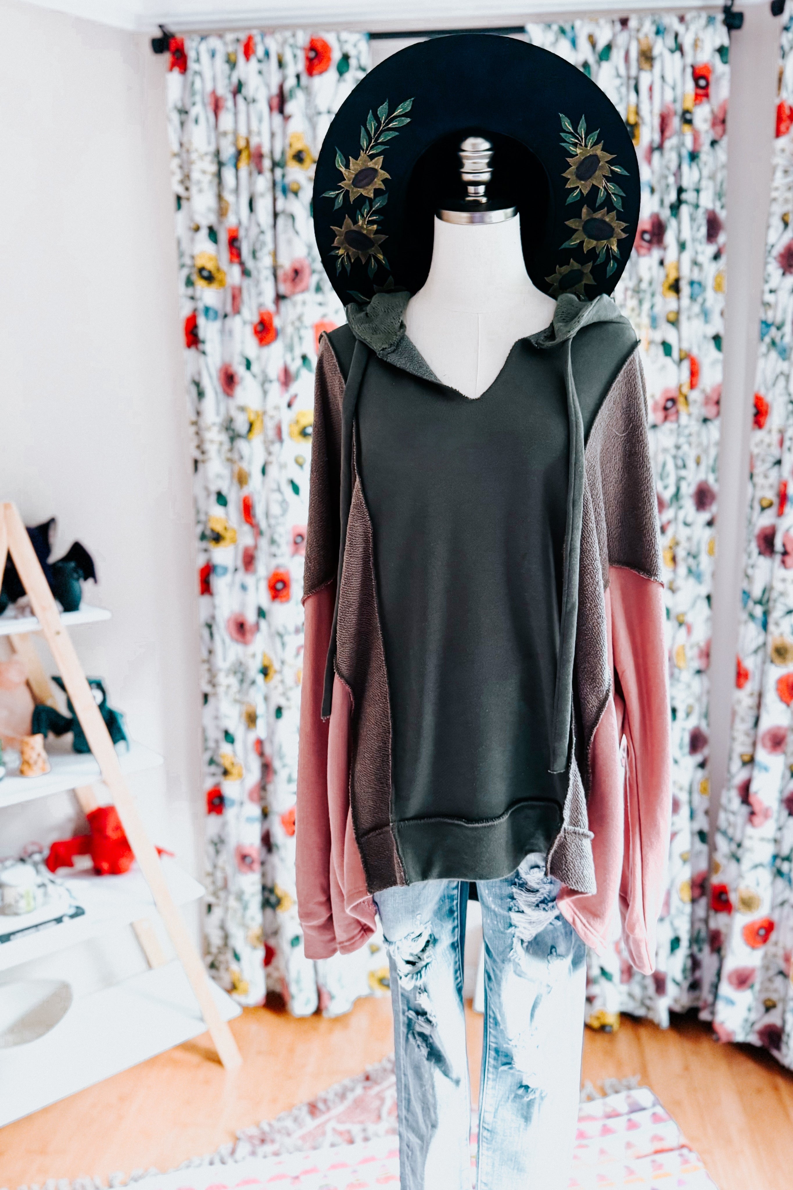 Mannequin dressed in a sage, muted brown, and dusty pink color block hoodie paired with acid-wash jeans and a black wide-brimmed hat with floral embroidery in a bright room.