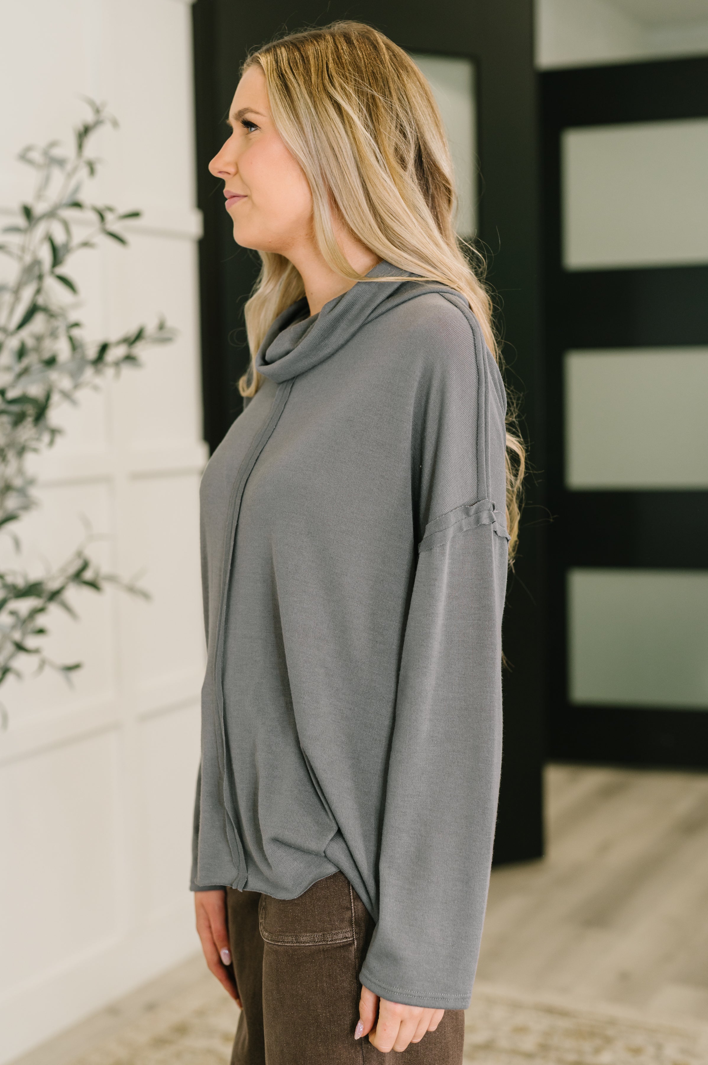 Profile view of a woman indoors wearing a relaxed grey turtleneck with a subtle center seam, paired with dark brown pants.