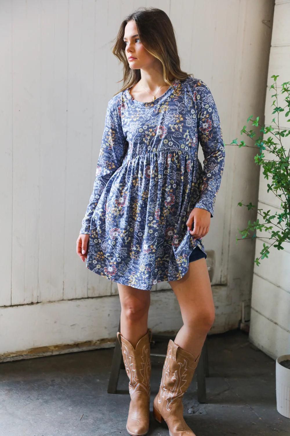 Woman wearing a blue floral patterned swing dress with long sleeves and a gathered waist, paired with tan embroidered cowboy boots indoors near a plant and white wall.