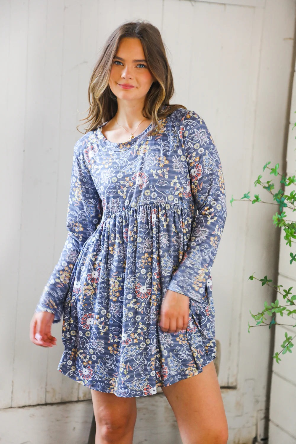 Young woman indoors wearing a blue floral patterned long-sleeve swing dress with a gathered waist, standing near a plant and white wooden wall.