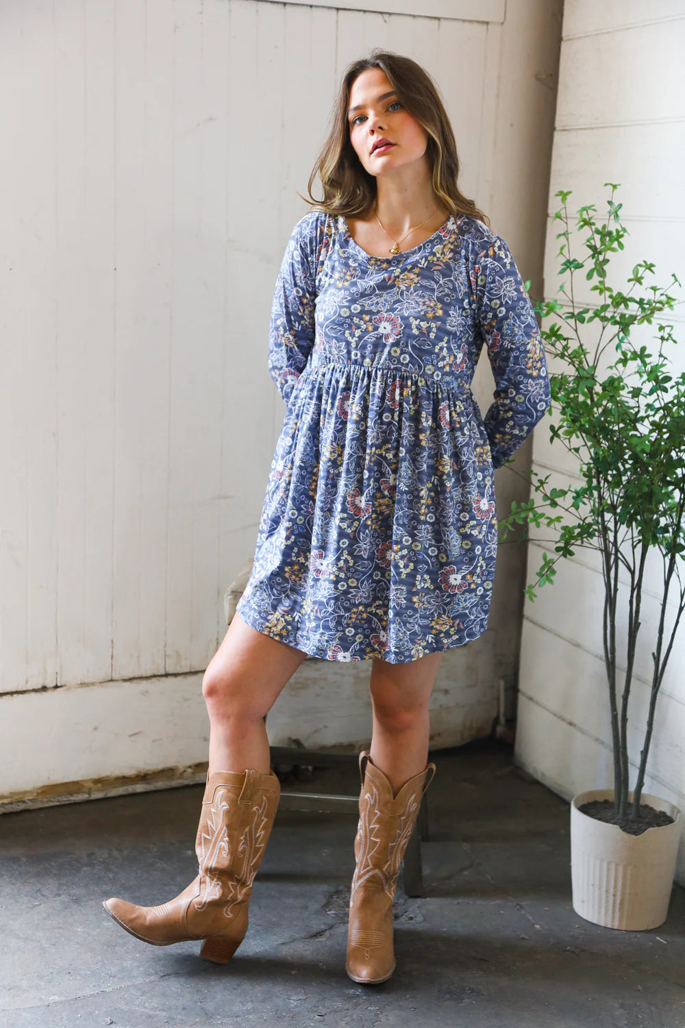 Woman standing indoors wearing a blue floral patterned swing dress with long sleeves, paired with tan embroidered cowboy boots near a potted plant.