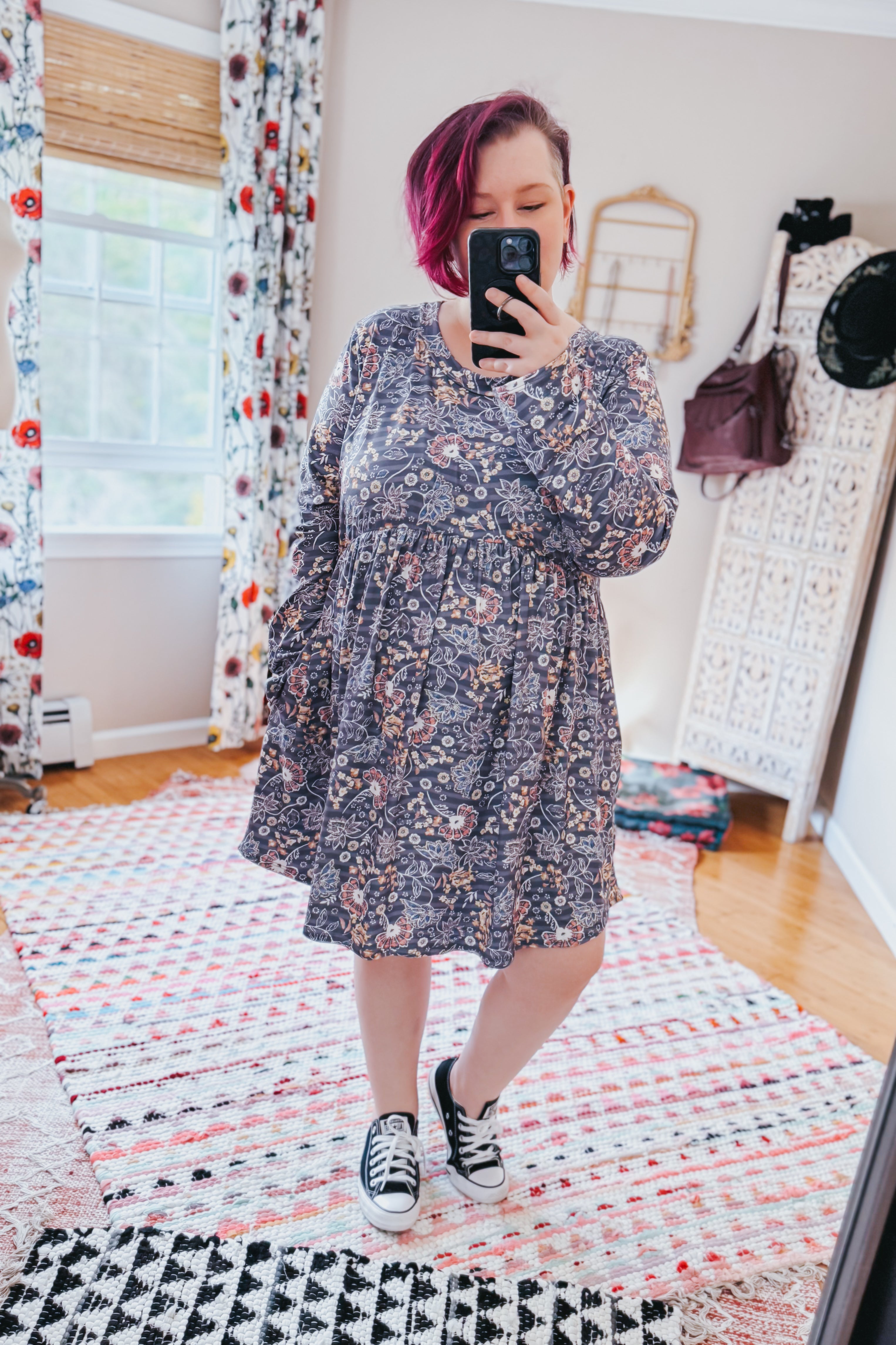 Woman wearing a blue floral patterned swing dress with long sleeves and an empire waist, paired with black sneakers in a bright room with patterned rugs and floral curtains.