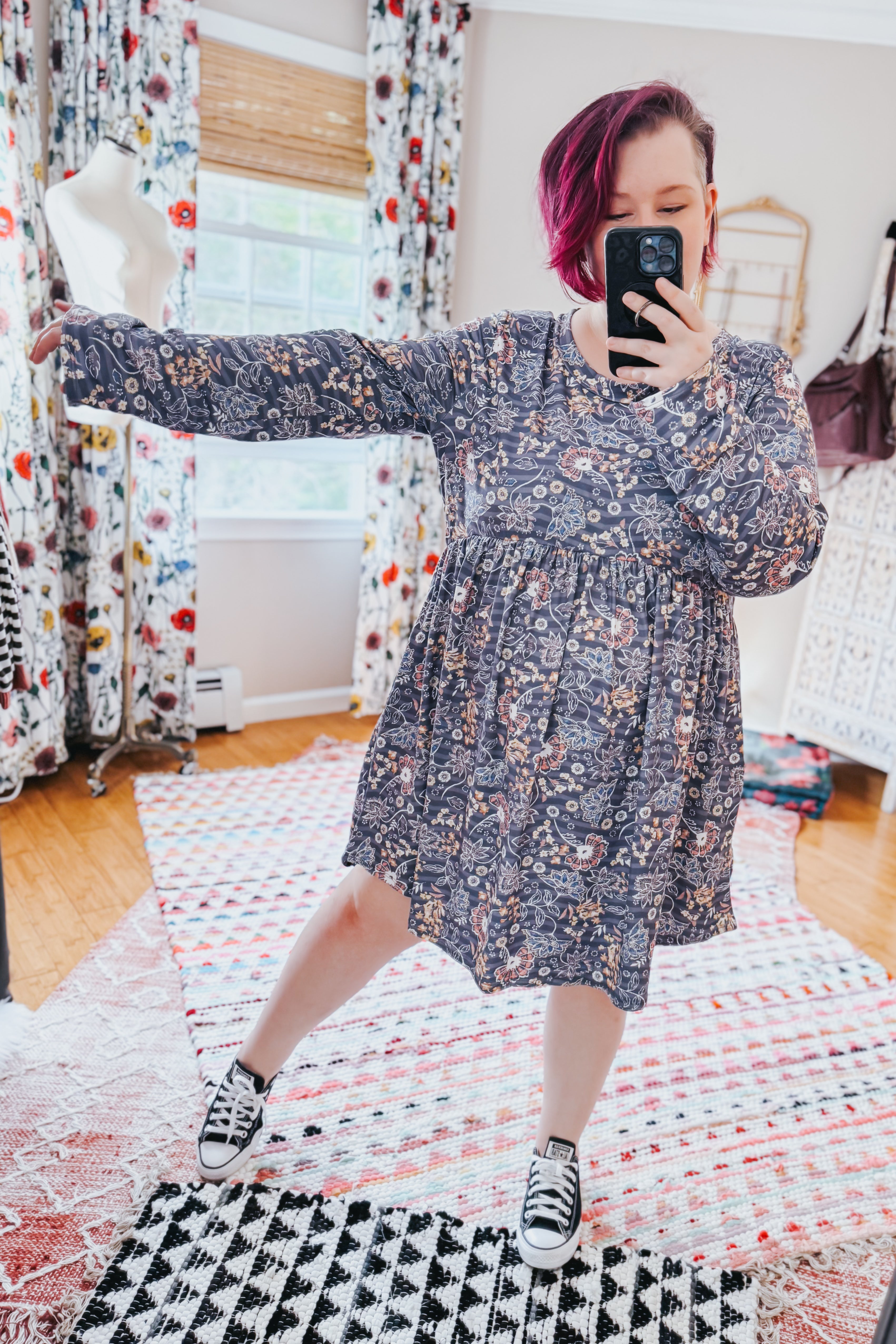 Woman in a blue floral patterned swing dress with long sleeves and gathered waist taking a selfie indoors on a vibrant multi-patterned rug.