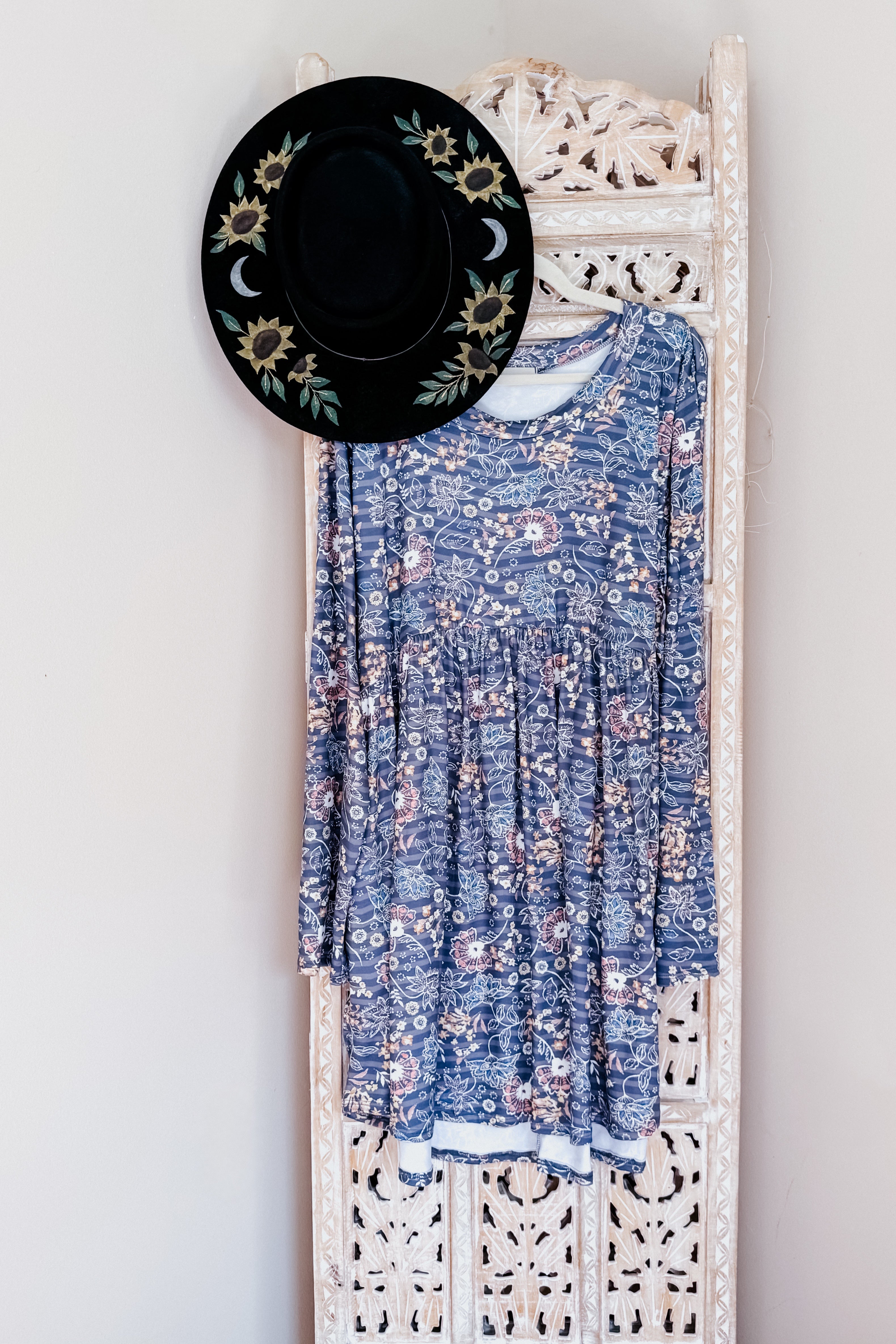 Blue long-sleeve swing dress with a floral print and gathered waist hanging on a carved wooden panel with a black wide-brimmed hat featuring embroidered sunflowers and crescent moons.