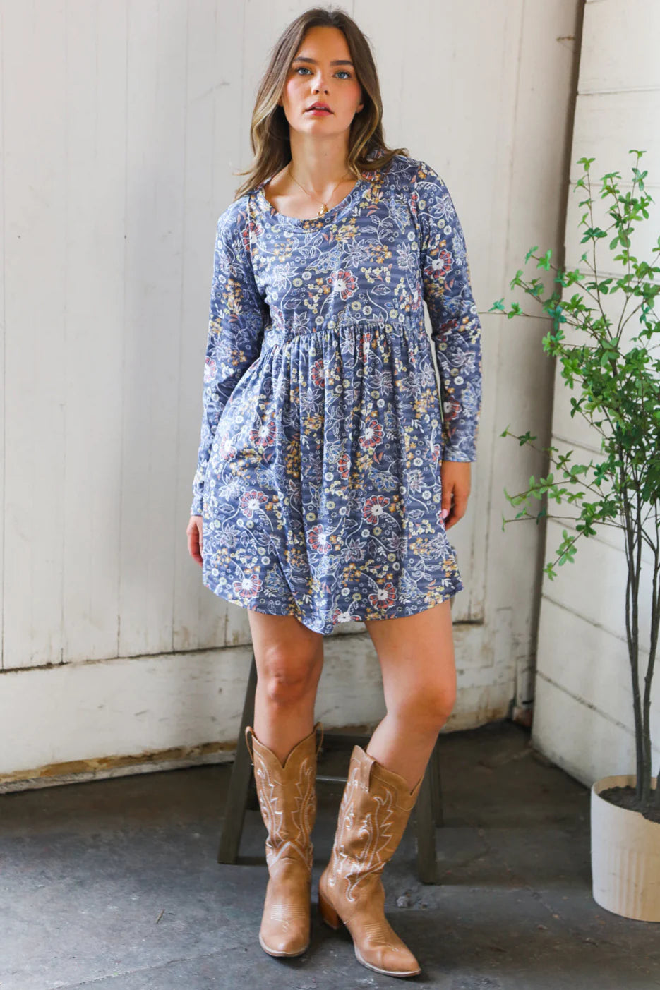 Young woman wearing a blue floral patterned swing dress with long sleeves and a gathered waist, paired with tan cowboy boots standing indoors near a potted plant.