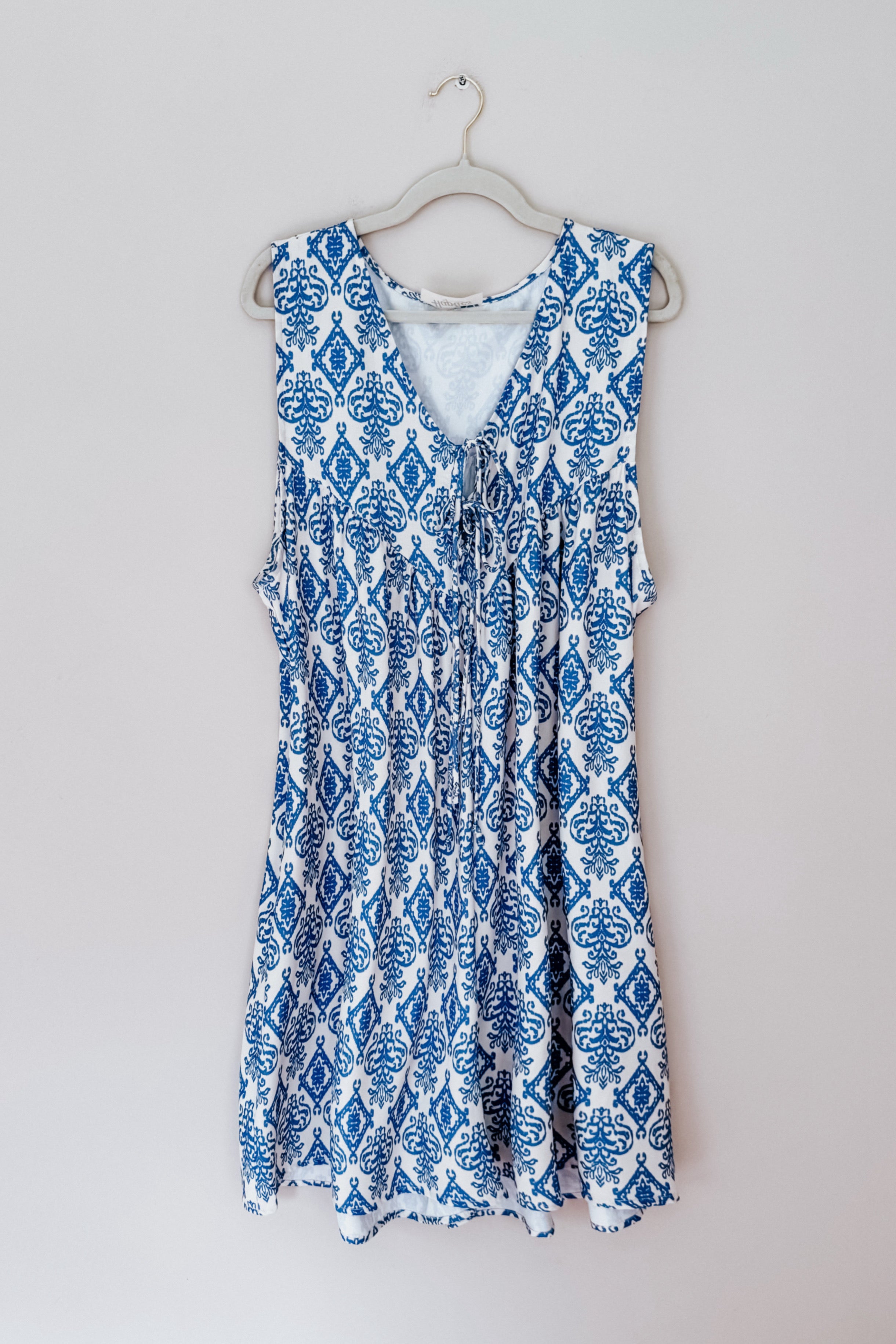 Sleeveless romper with a deep V-neck and adjustable tie, featuring an intricate blue pattern on a white background hanging on a white hanger against a plain wall.