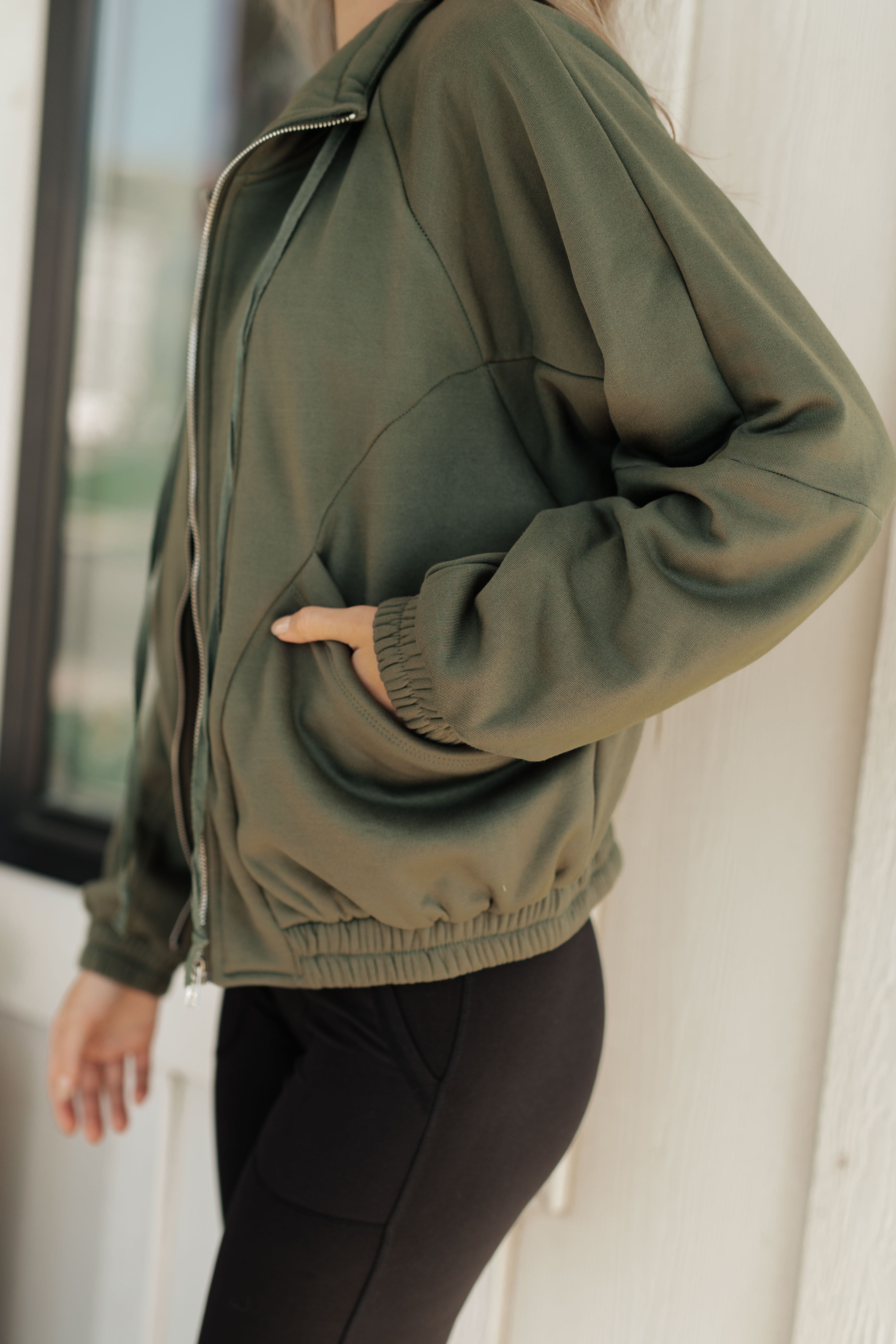 Person wearing an olive green zip-up sweatshirt with elastic cuffs and hem, featuring side pockets, paired with black leggings, standing near a window.