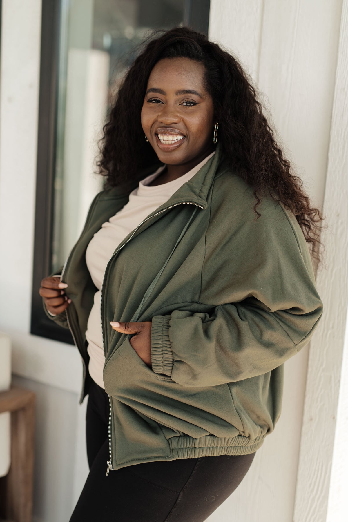 Woman modeling an olive zip front sweatshirt with a high stand collar and gathered sleeves over a cream top indoors