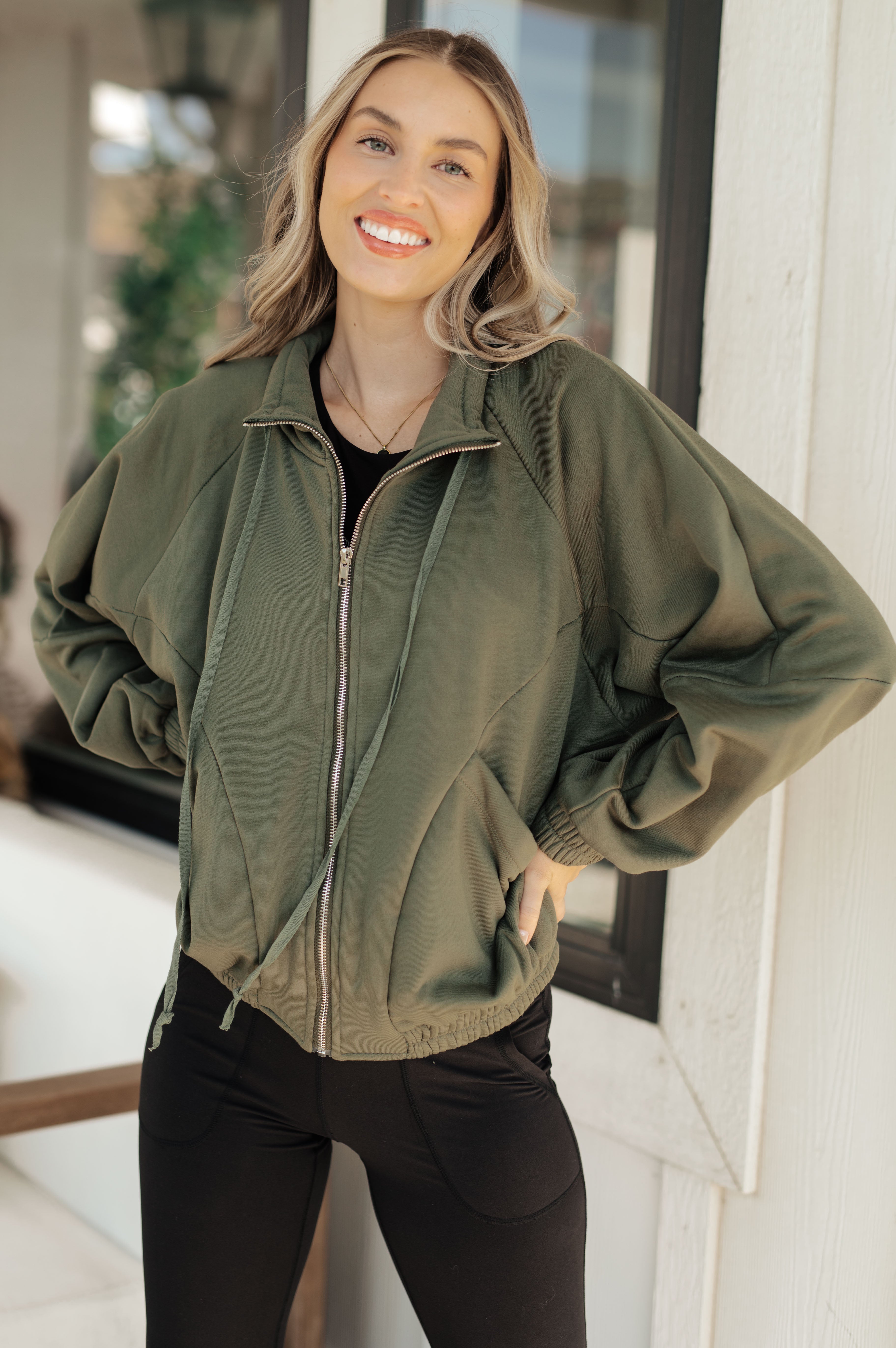 Smiling woman wearing an olive zip-up sweatshirt with loose sleeves and elastic cuffs, paired with black pants, standing indoors by a window.