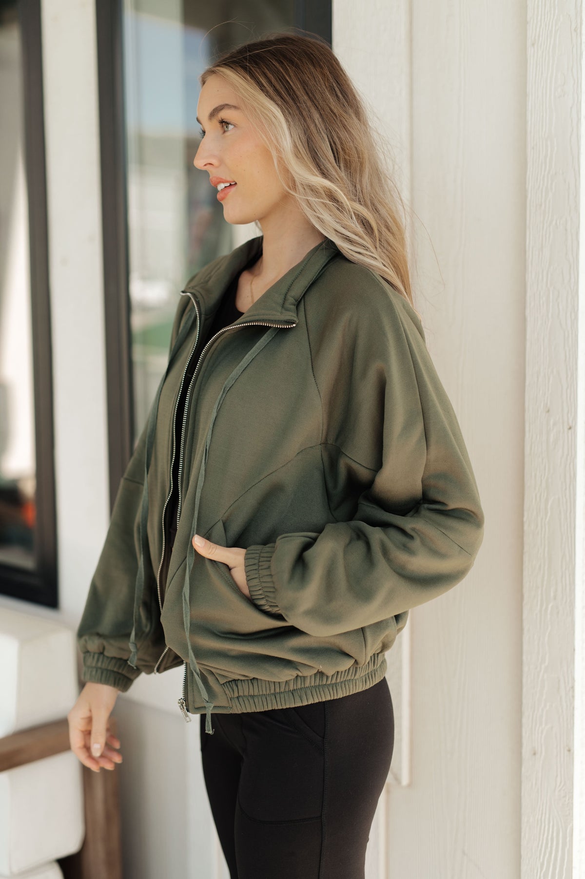 Woman in an olive zip front sweatshirt with a high stand collar and snap pockets poses in profile with black pants indoors