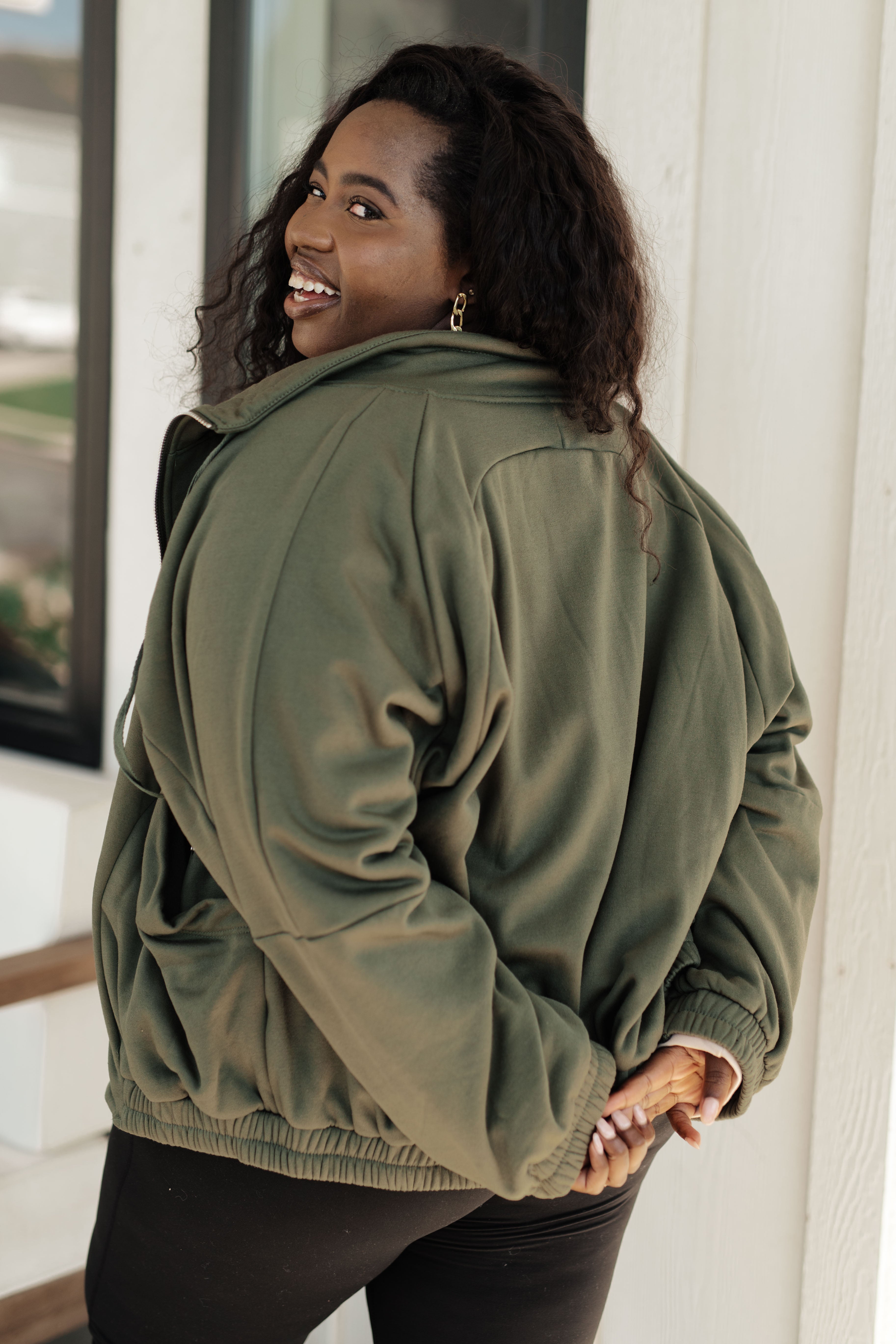 Happy woman wearing an oversized olive zip-up sweatshirt with elastic cuffs and hem, standing outdoors near a white wall.
