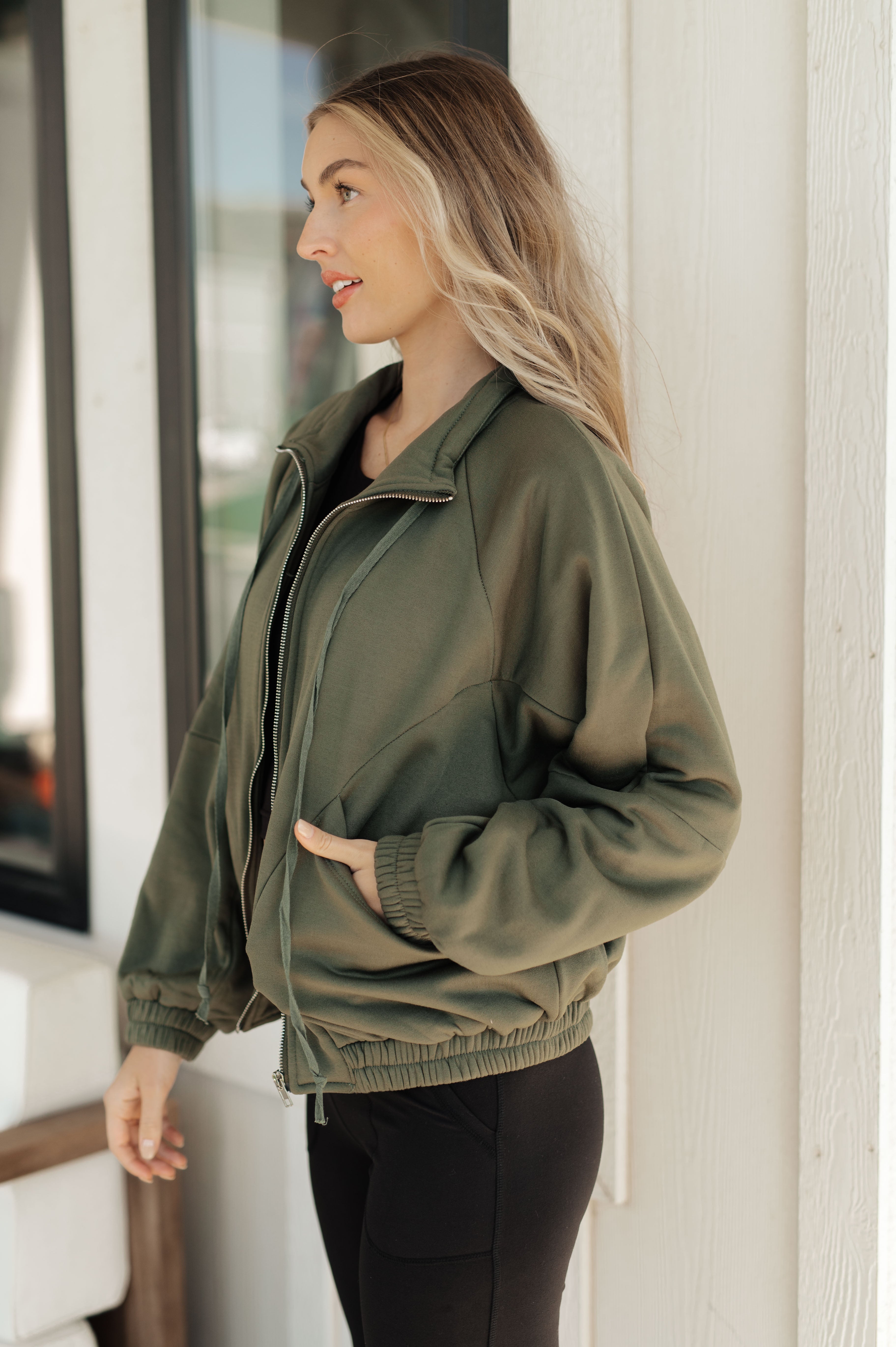 Woman wearing an olive zip-up sweatshirt with elastic cuffs and hem, featuring a relaxed fit and side pockets, standing indoors by a window.