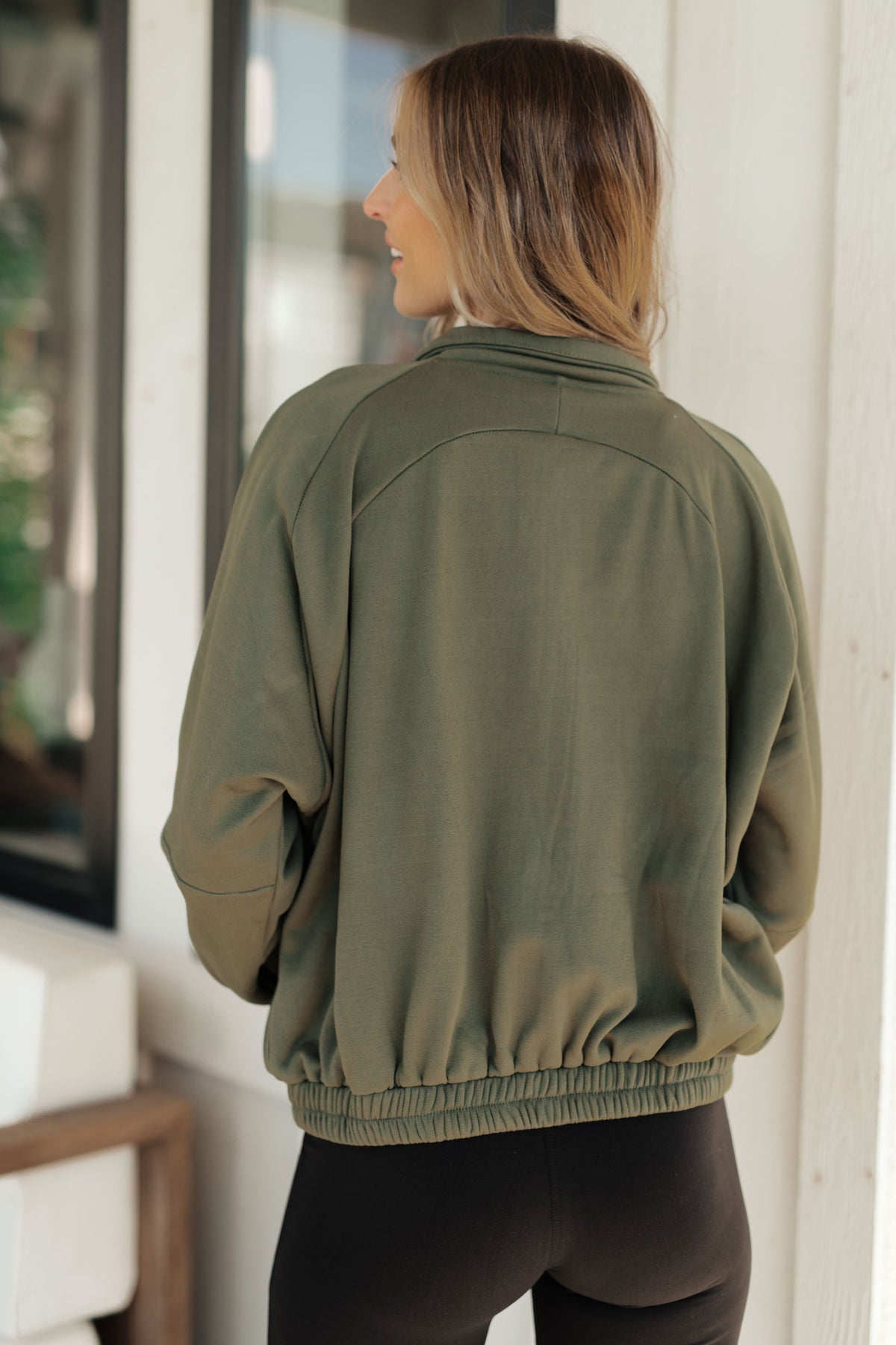Back view of a woman wearing an olive sweatshirt with a stand collar gathered sleeves and elastic hem paired with black pants indoors