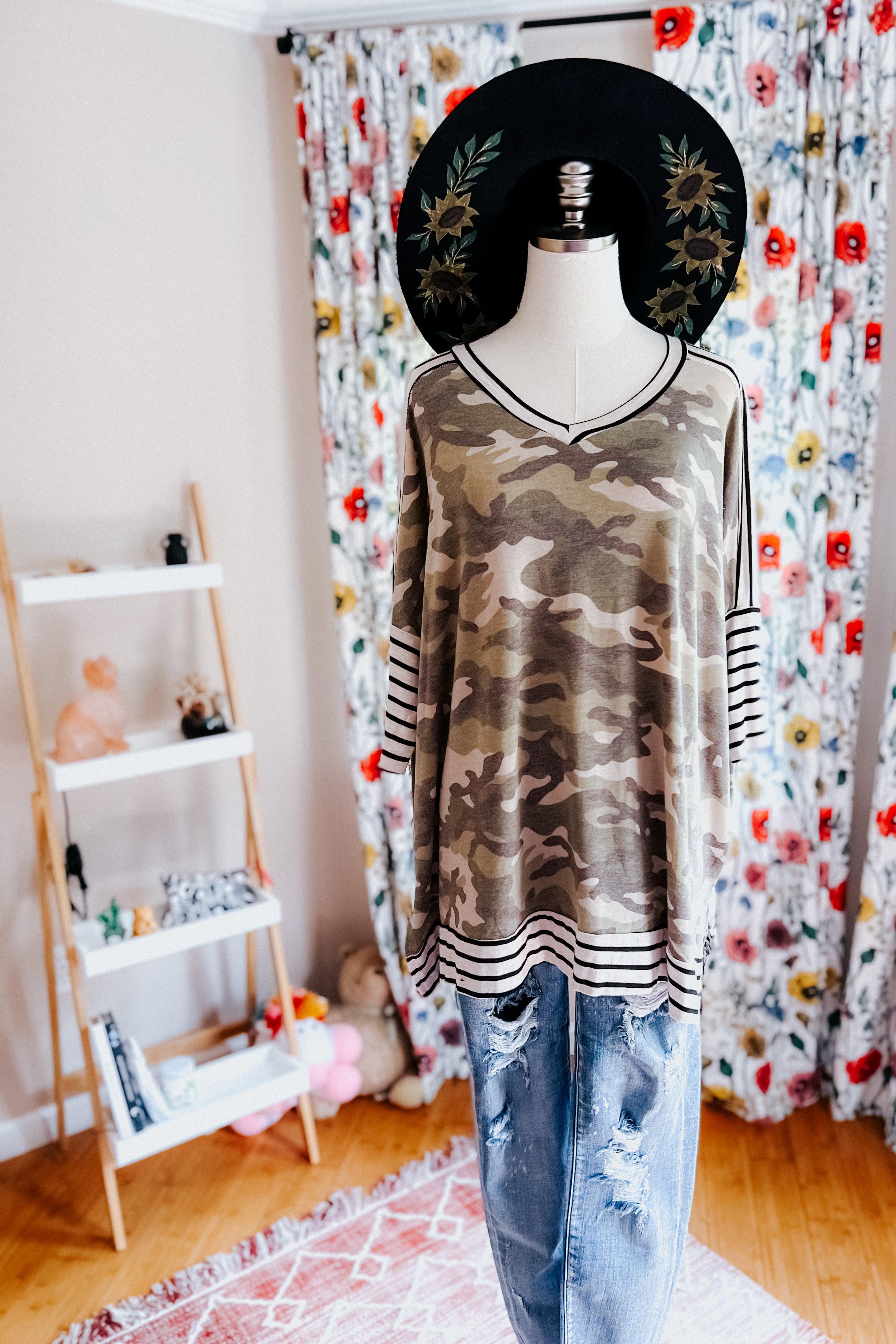 Loose-fitting green camo patterned tunic with striped mid sleeves and hem paired with distressed blue jeans on a mannequin with a black floral hat