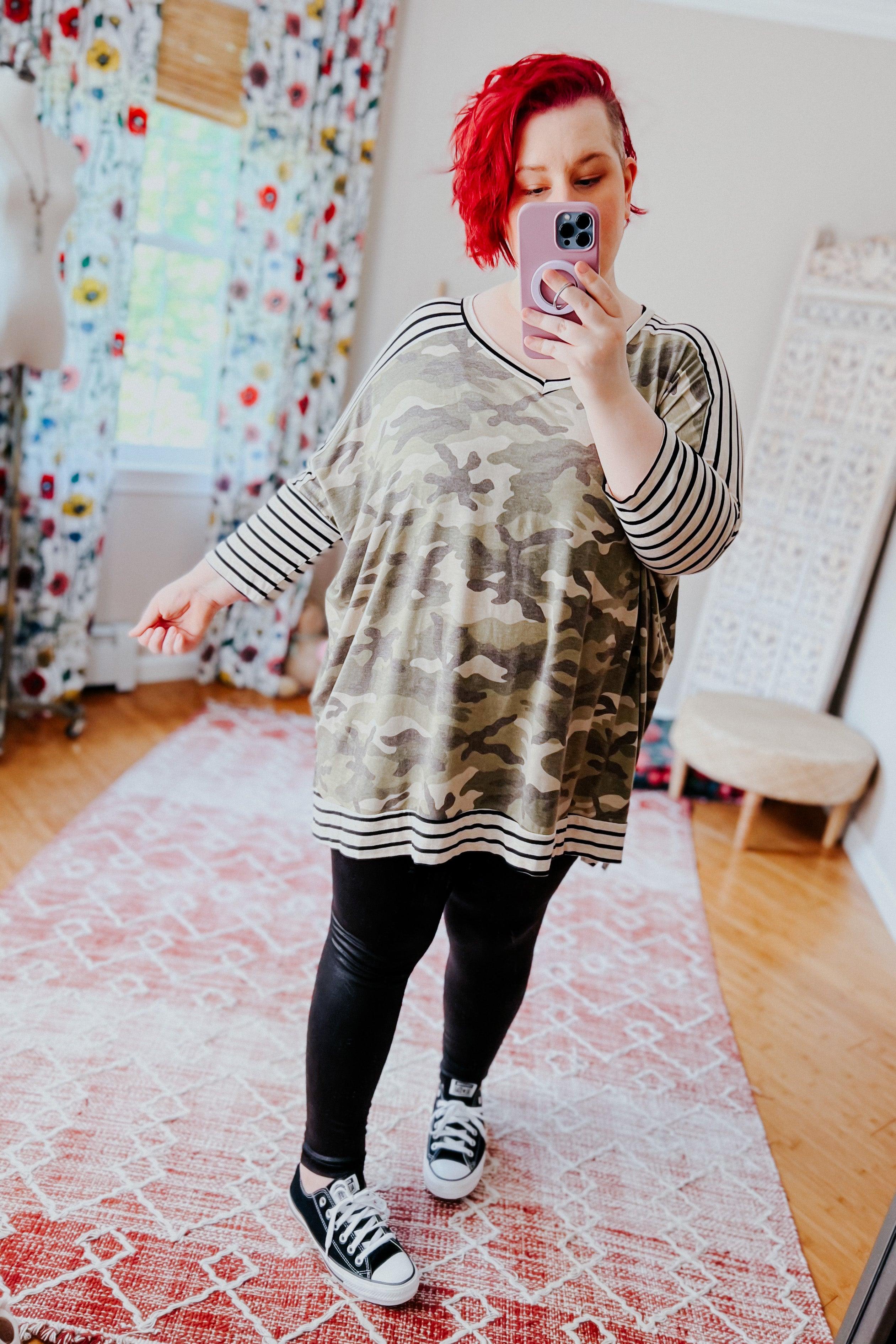 Woman with short red hair wearing an oversized green camo patterned tunic with black and white striped mid sleeves, neckline, and hem, paired with black leggings and black sneakers, taking a mirror selfie in a bright room with floral curtains and light wood flooring.