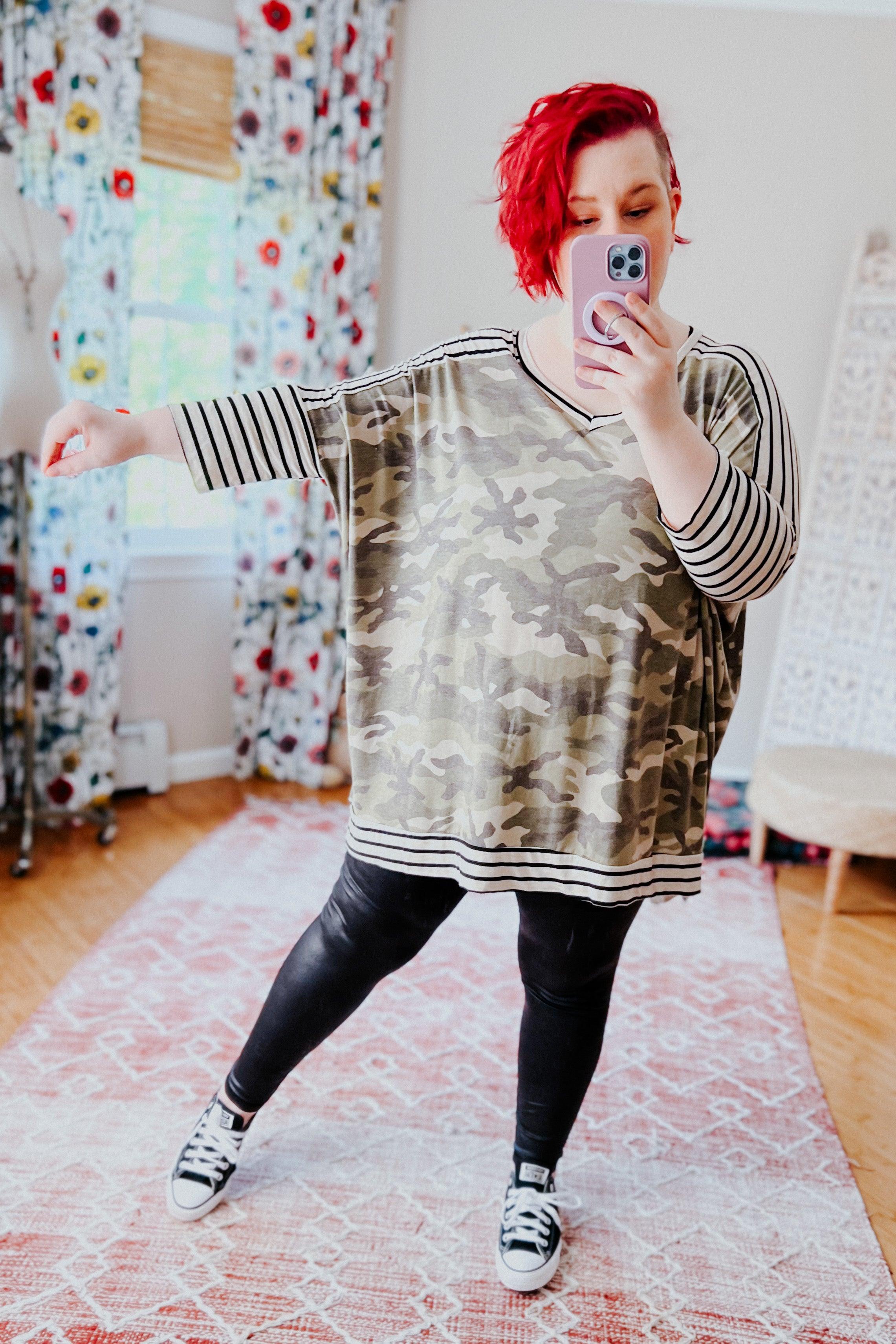 Plus-size woman with short red hair wearing a loose green camo patterned mid sleeve tunic with striped sleeves and hem, paired with black leggings and sneakers, taking a mirror selfie in a bright room with floral curtains and light hardwood floors.