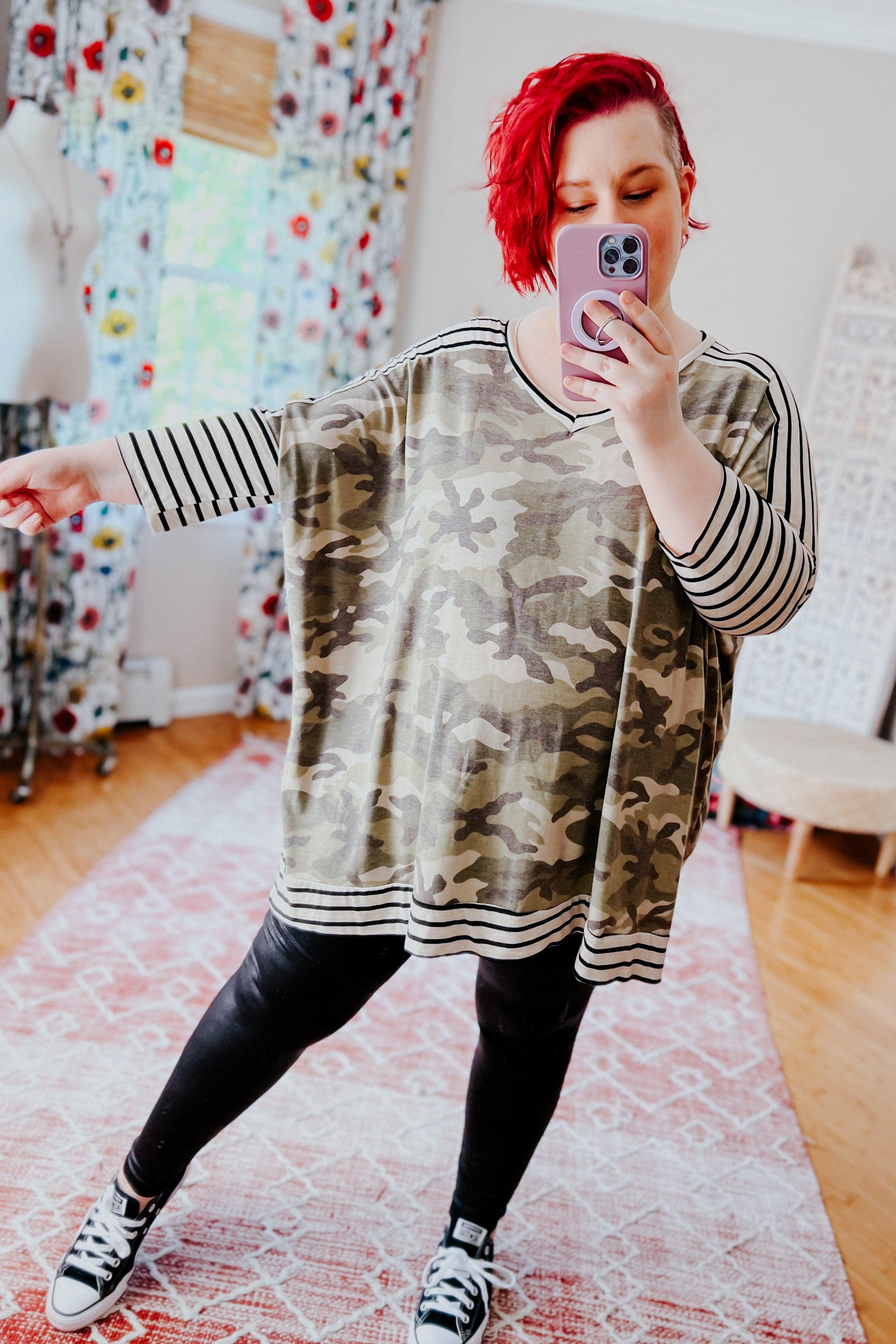 Woman with short red hair wearing a loose green camo patterned mid sleeve tunic with contrasting black and white striped sleeves, neckline, and hem, paired with black leggings and sneakers, taking a mirror selfie in a warmly lit room with patterned curtains and a pink area rug.