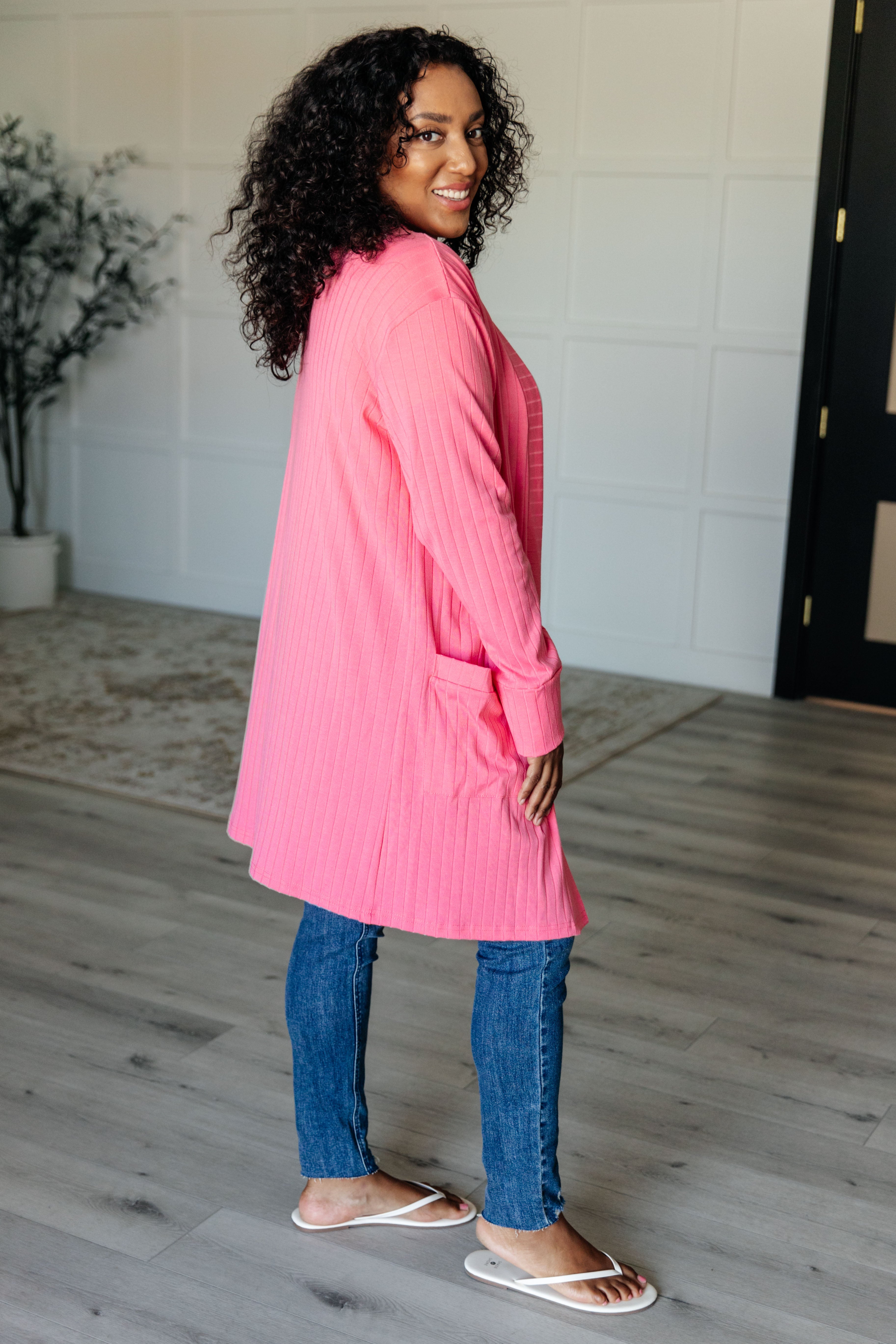 Woman smiling over shoulder wearing bright pink ribbed Modern Idealist open front cardigan with pockets standing indoors