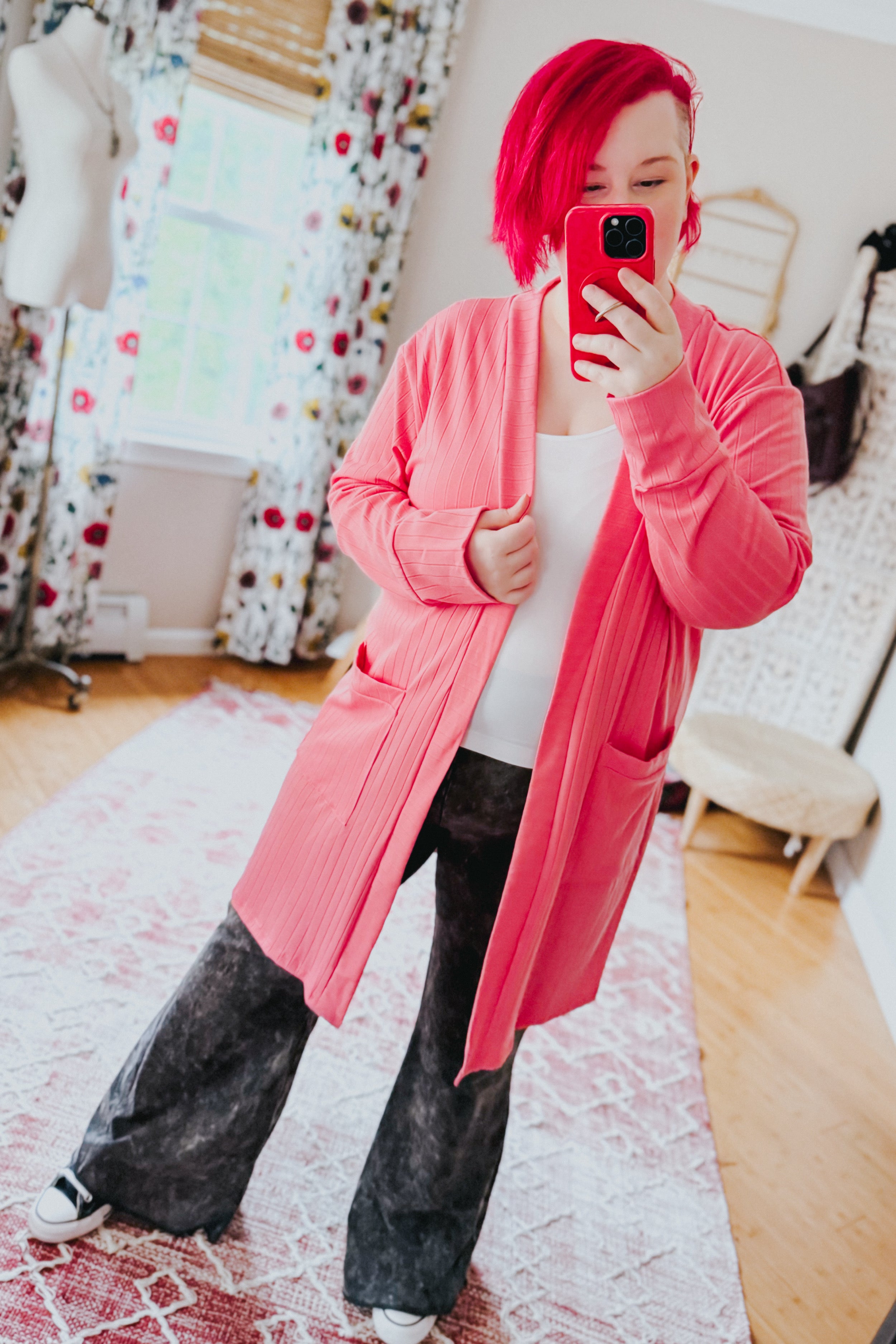 Woman taking mirror selfie wearing coral ribbed Modern Idealist open front cardigan with pockets over white top and wide dark pants indoors