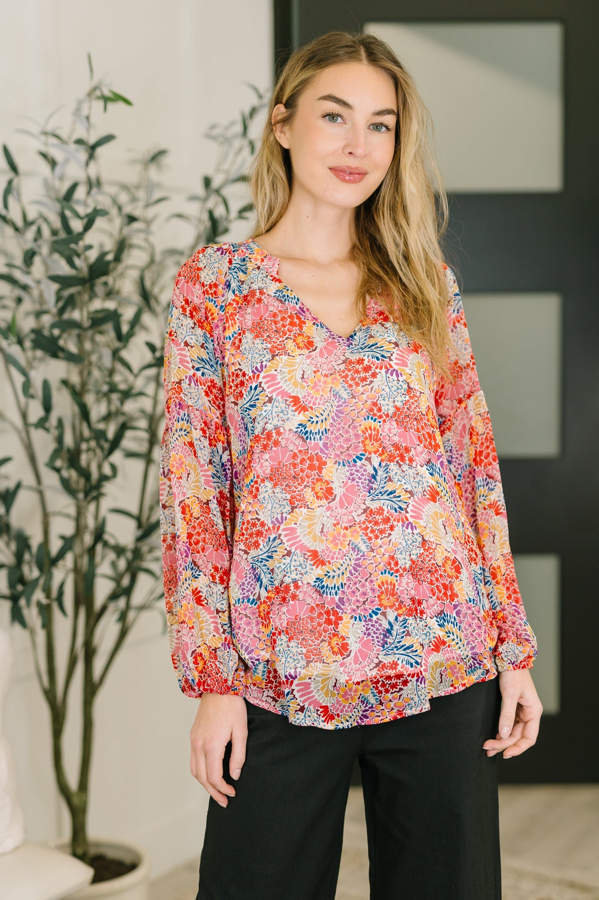 Woman modeling a pink floral V neck blouse with voluminous sleeves and black pants in a bright indoor setting