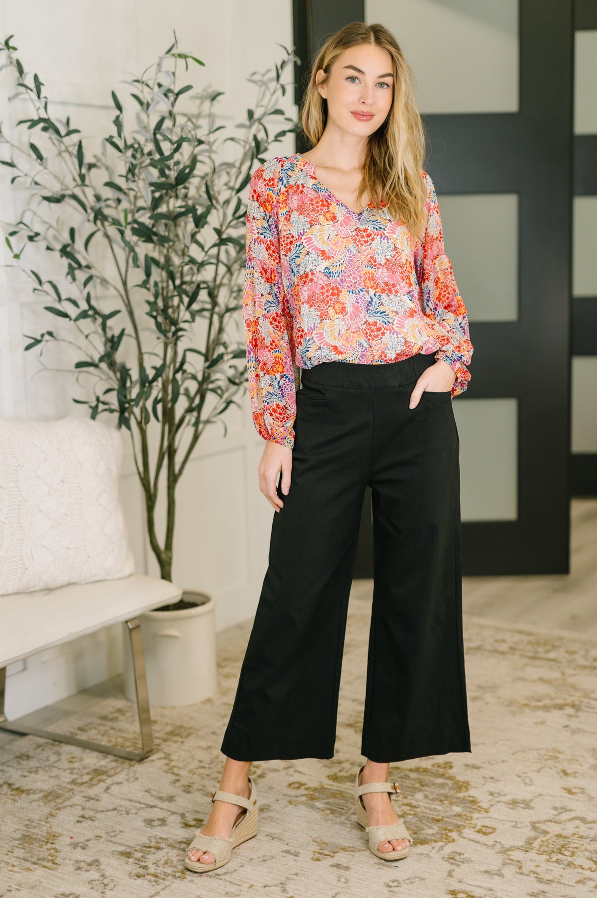 Woman modeling a pink floral V neck blouse with puff sleeves black wide leg pants and strappy sandals in a stylish indoor setting