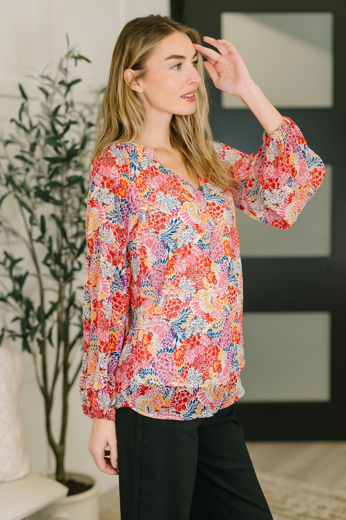 Woman wearing a pink floral V-neck blouse with dramatic puff sleeves and black trousers in a softly lit home interior