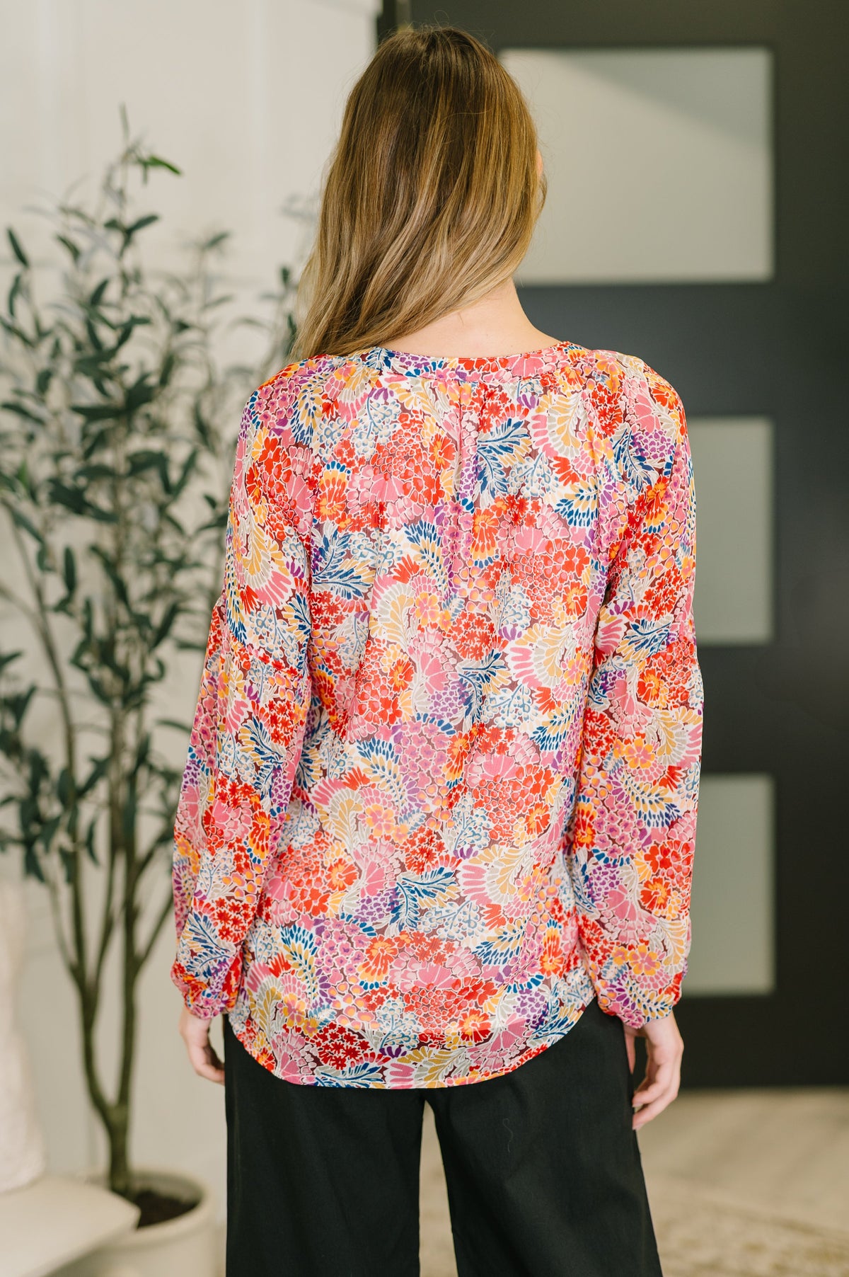 Woman viewed from behind wearing a vibrant pink floral blouse with long puff sleeves and black pants in a bright indoor setting