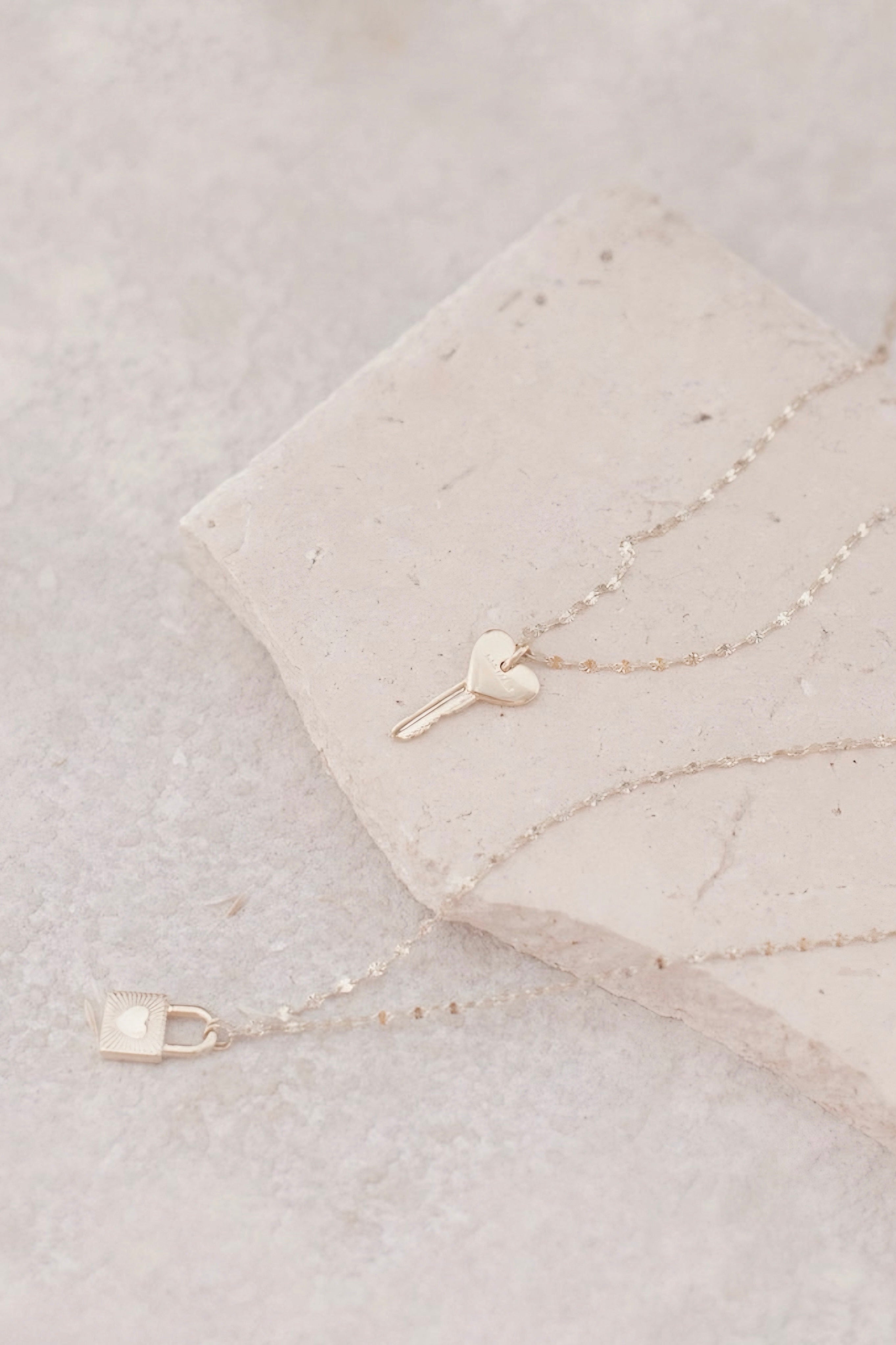 Delicate gold-toned necklace featuring a heart-shaped key pendant and a small padlock charm on a fine chain, styled on a light stone surface.