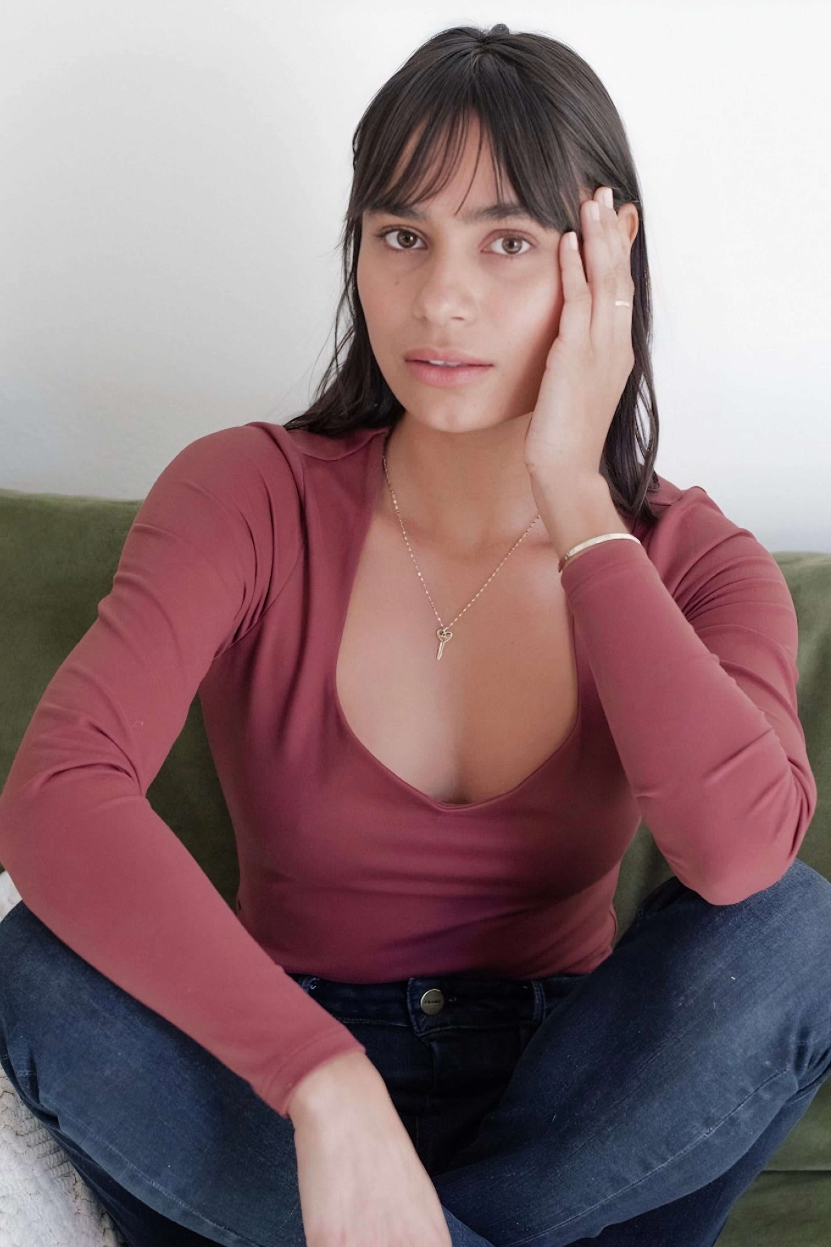 Woman sitting cross-legged on a green sofa wearing a long-sleeve mauve top with a delicate gold key necklace and dark jeans.