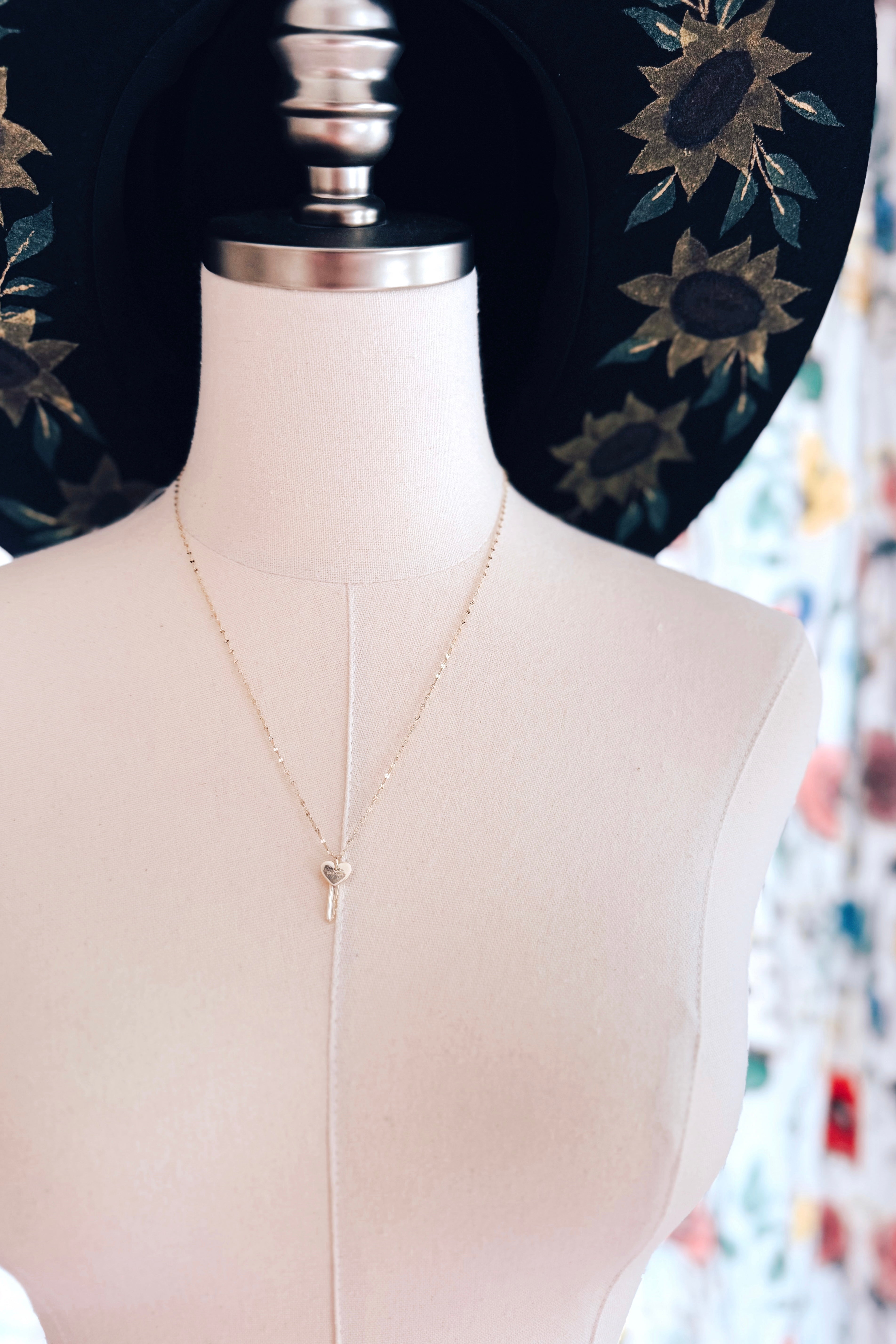 Gold delicate key pendant necklace with a heart-shaped top displayed on a white dress form with a decorative black hat featuring floral details in the background.