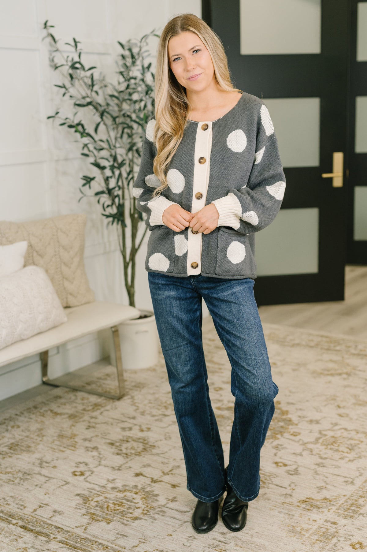 Woman wearing a charcoal button front cardigan with oversized cream polka dots, jeans, and black shoes indoors