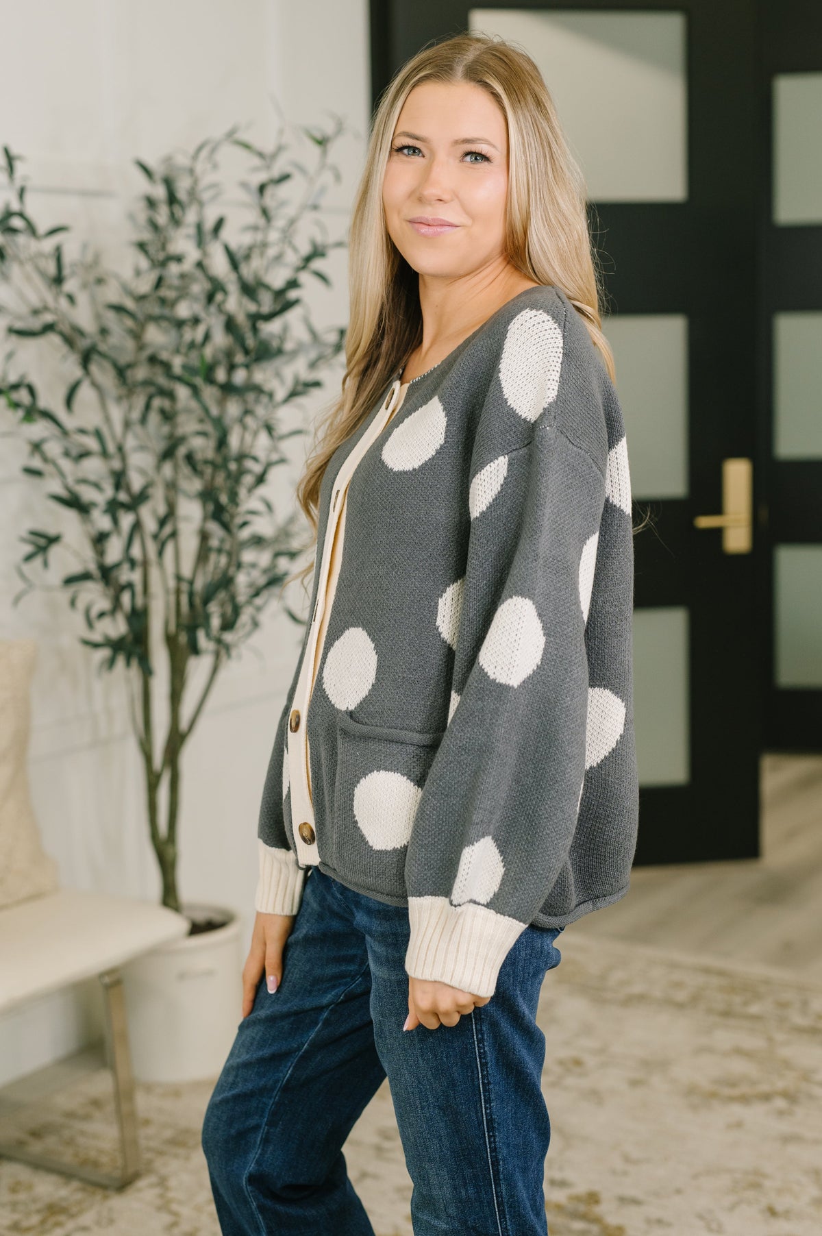 Woman wearing a charcoal knit button down cardigan with large white polka dots and ribbed cuffs paired with jeans indoors