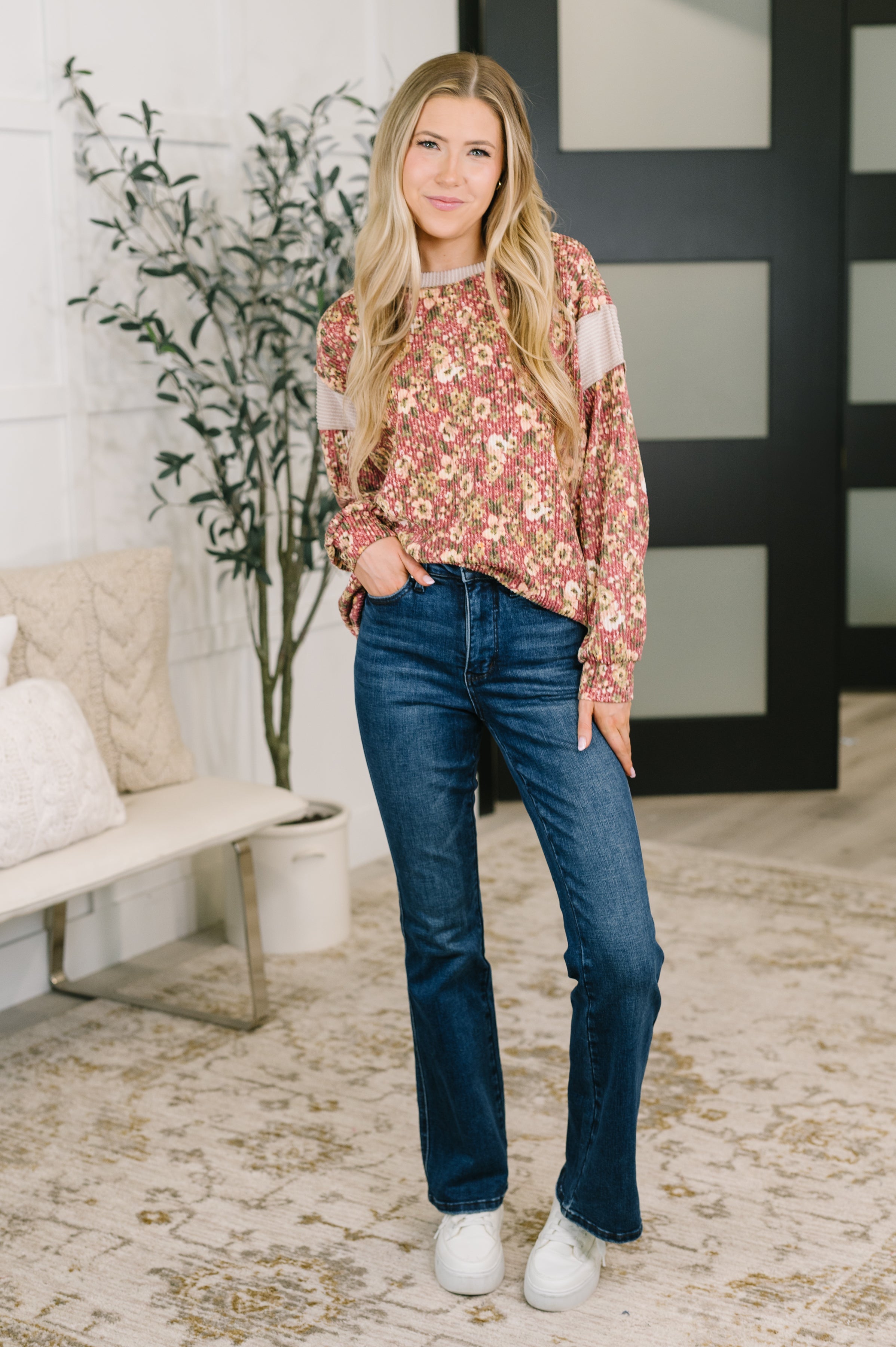Woman wearing a rust floral textured long sleeve top with relaxed sleeves dark jeans and white sneakers indoors