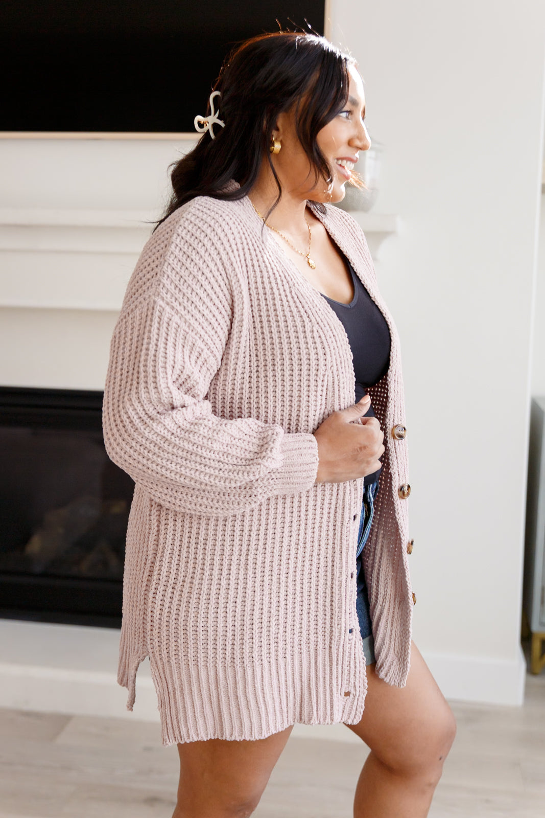 Woman wearing a light pink button down cardigan over a black top and blue jeans shorts standing in an indoor setting.