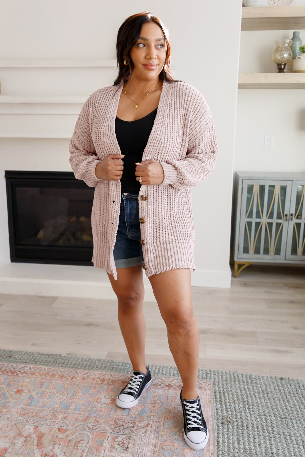 Woman wearing a light pink button down cardigan over a black top, blue jeans shorts and black converse sneakers standing in an indoor setting.