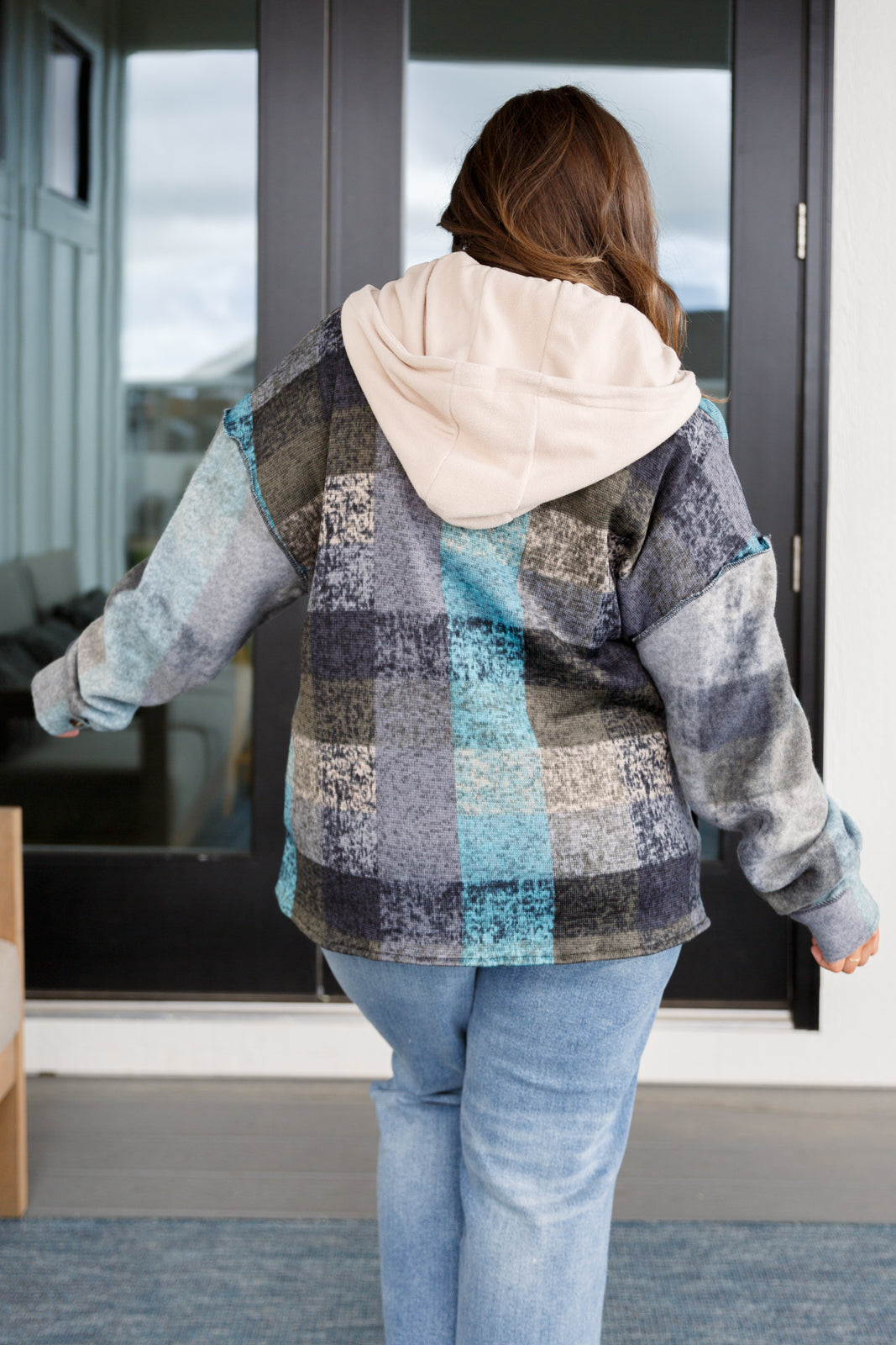 Woman seen from behind wearing a blue and gray plaid hooded shacket layered over a light beige hoodie, paired with light blue jeans.