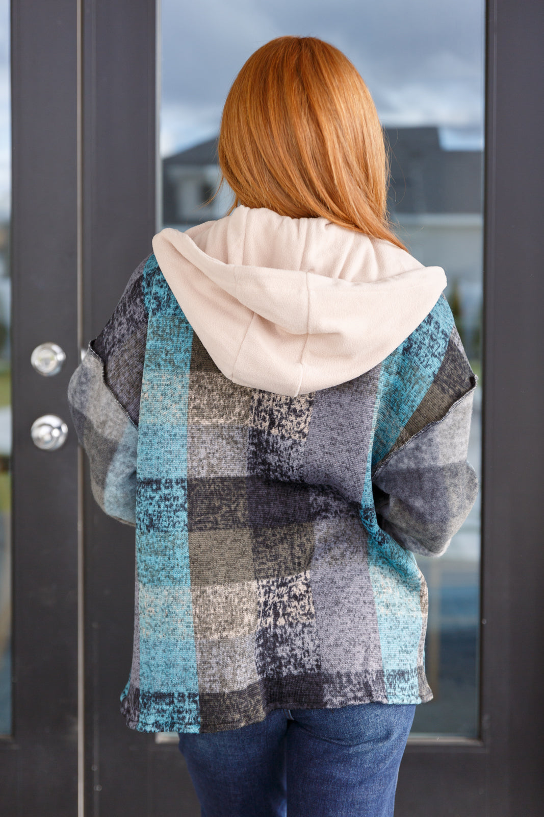 Woman seen from behind wearing a blue patchwork hooded shacket with a cream-colored hood, paired with dark blue jeans.