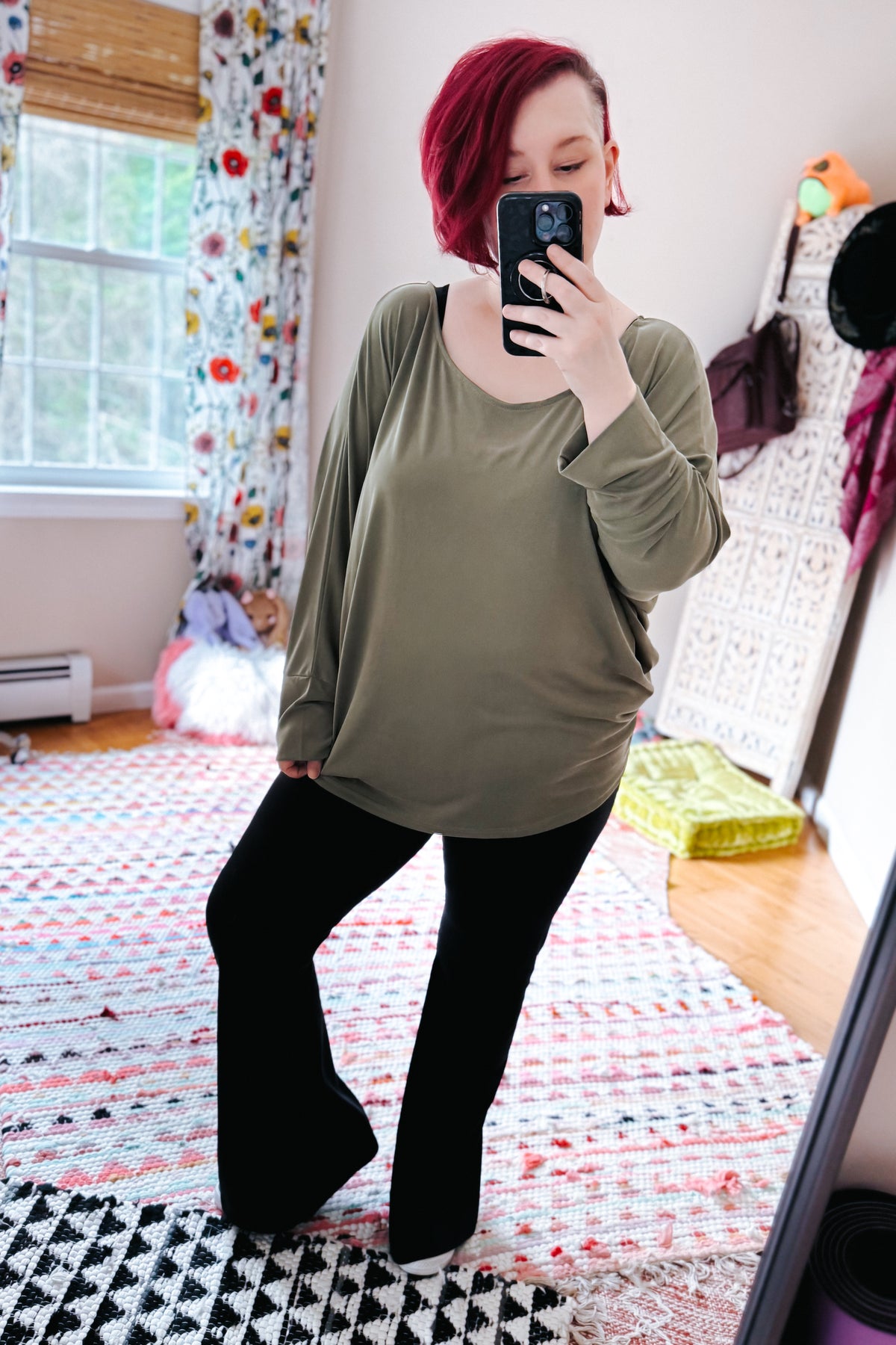 Red haired woman taking a mirror selfie in a green boat neck top with dolman sleeves and black pants in a colorful bedroom