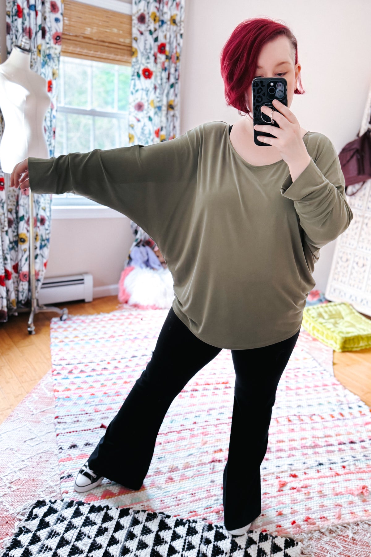 Woman taking a mirror selfie in a green boat neck top with dolman sleeves and exaggerated cuffs black flared pants and sneakers in a bedroom