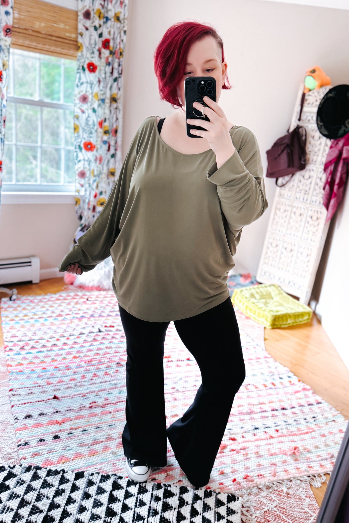 Red haired woman taking a mirror selfie in a green boat neck top with dolman sleeves black flared pants and sneakers in a bedroom
