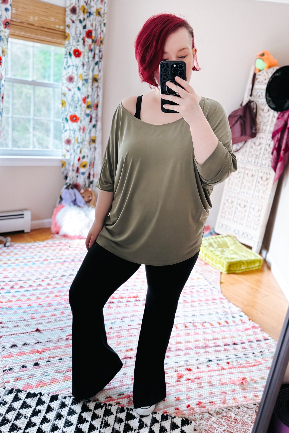Red haired woman taking a bedroom mirror selfie in a green boat neck top with dolman sleeves and black flared pants