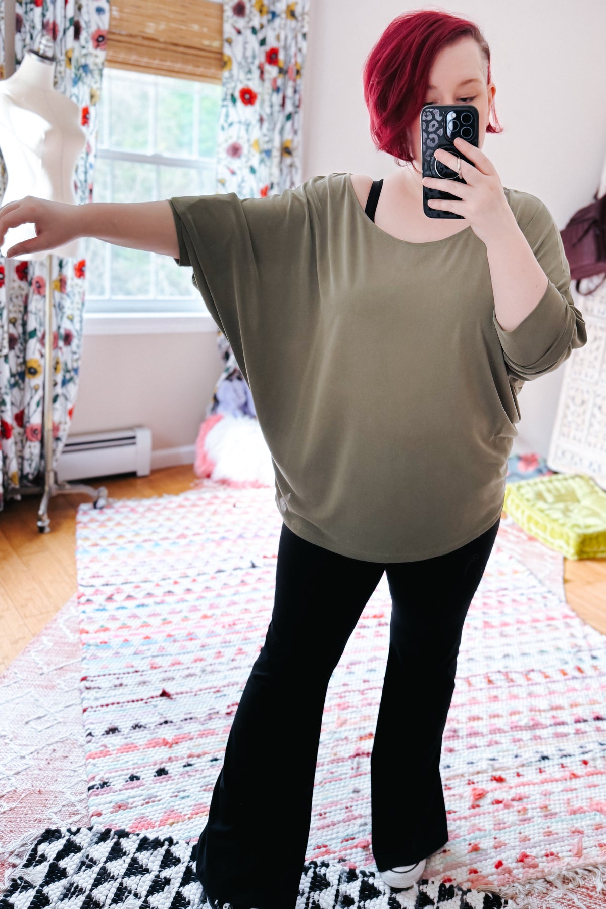 Red haired woman taking a bedroom mirror selfie in a green boat neckline top with dolman sleeves and black flared pants beside a colorful rug