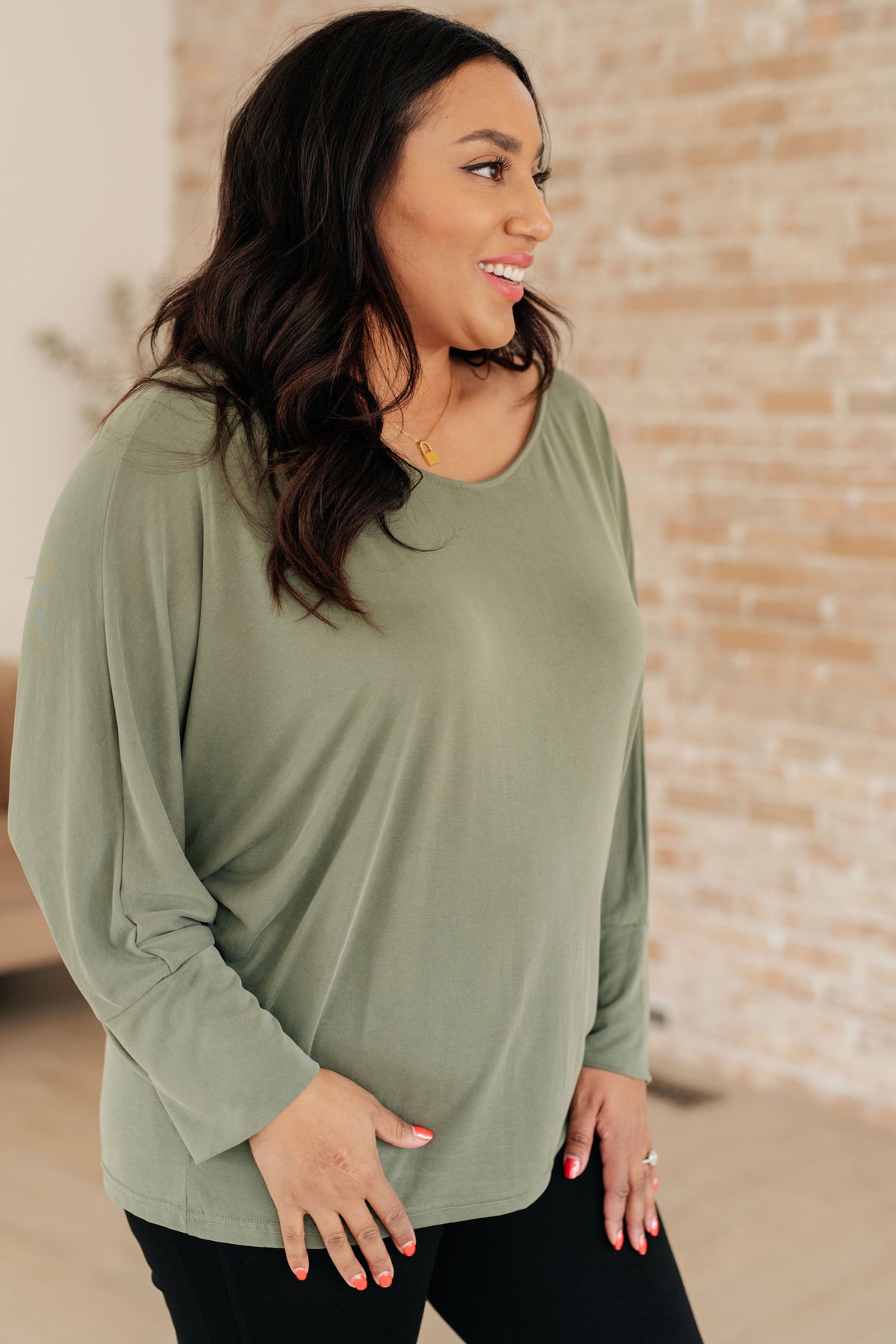 Smiling woman wearing a green boat neck long sleeve top with relaxed dolman sleeves and black pants in a bright brick walled room