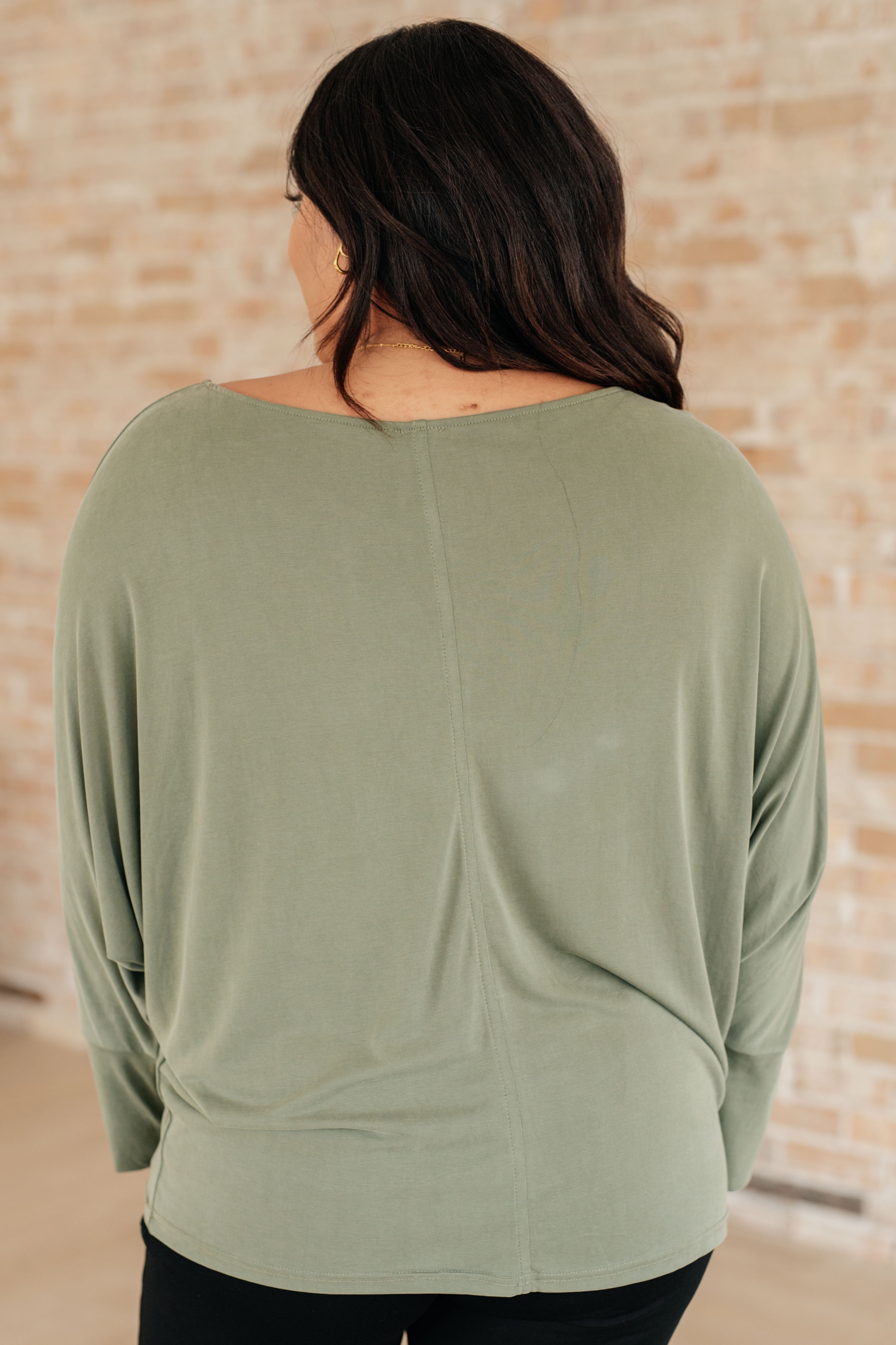 Back view of a woman wearing a green boat neck top with relaxed dolman sleeves and extended cuffs over black pants indoors