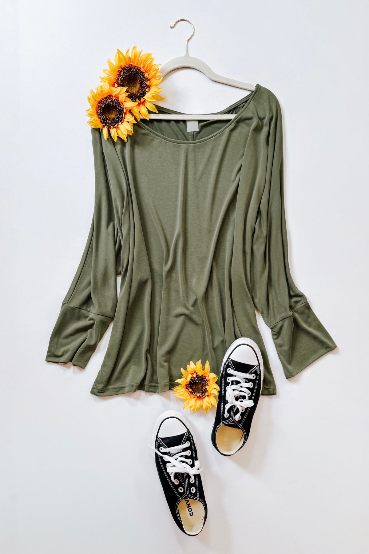 Green boat neck long sleeve top with dolman sleeves and wide cuffs displayed on a hanger with sunflowers and black sneakers
