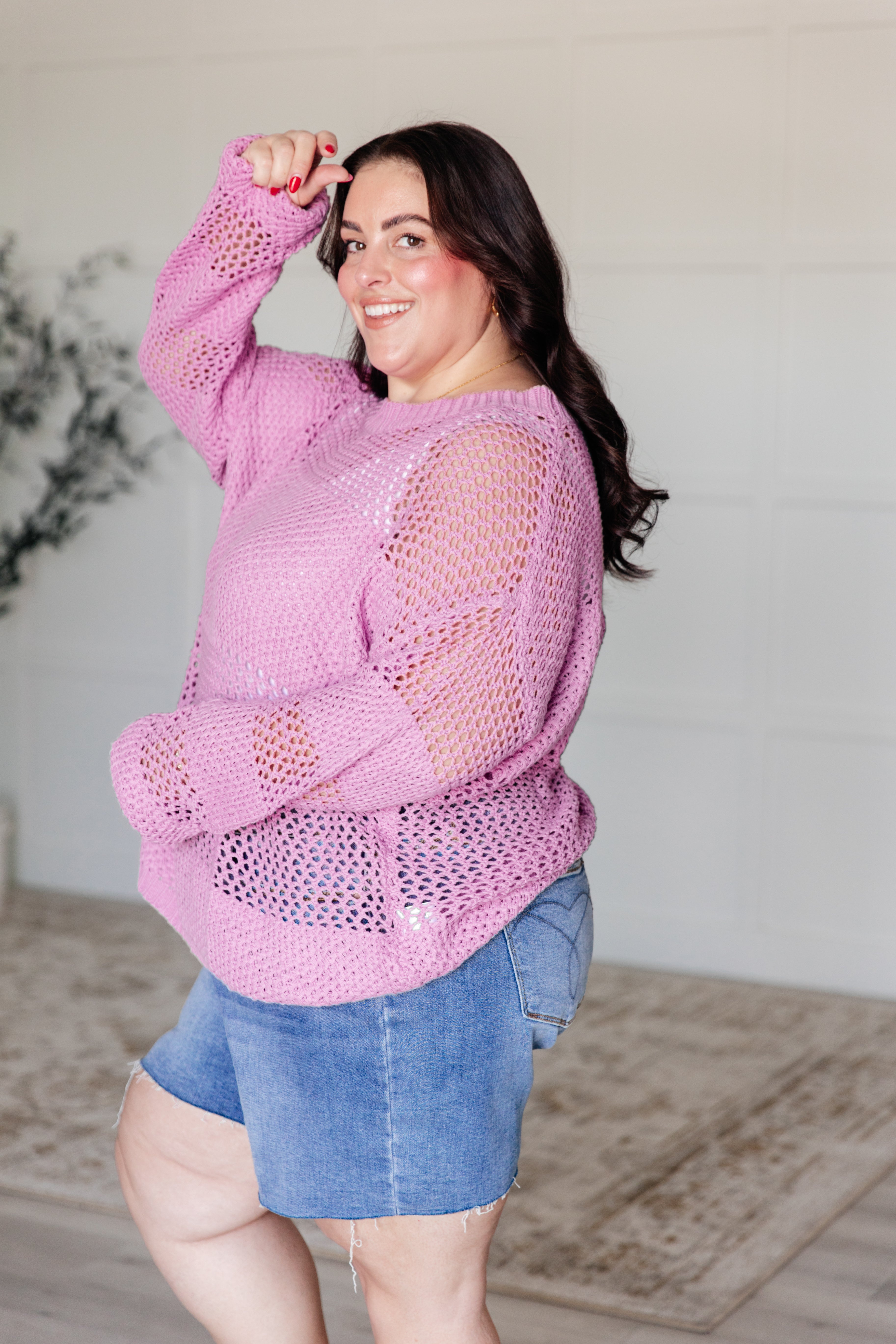 Plus-size woman smiling wearing a loose knit pink sweater paired with denim shorts indoors