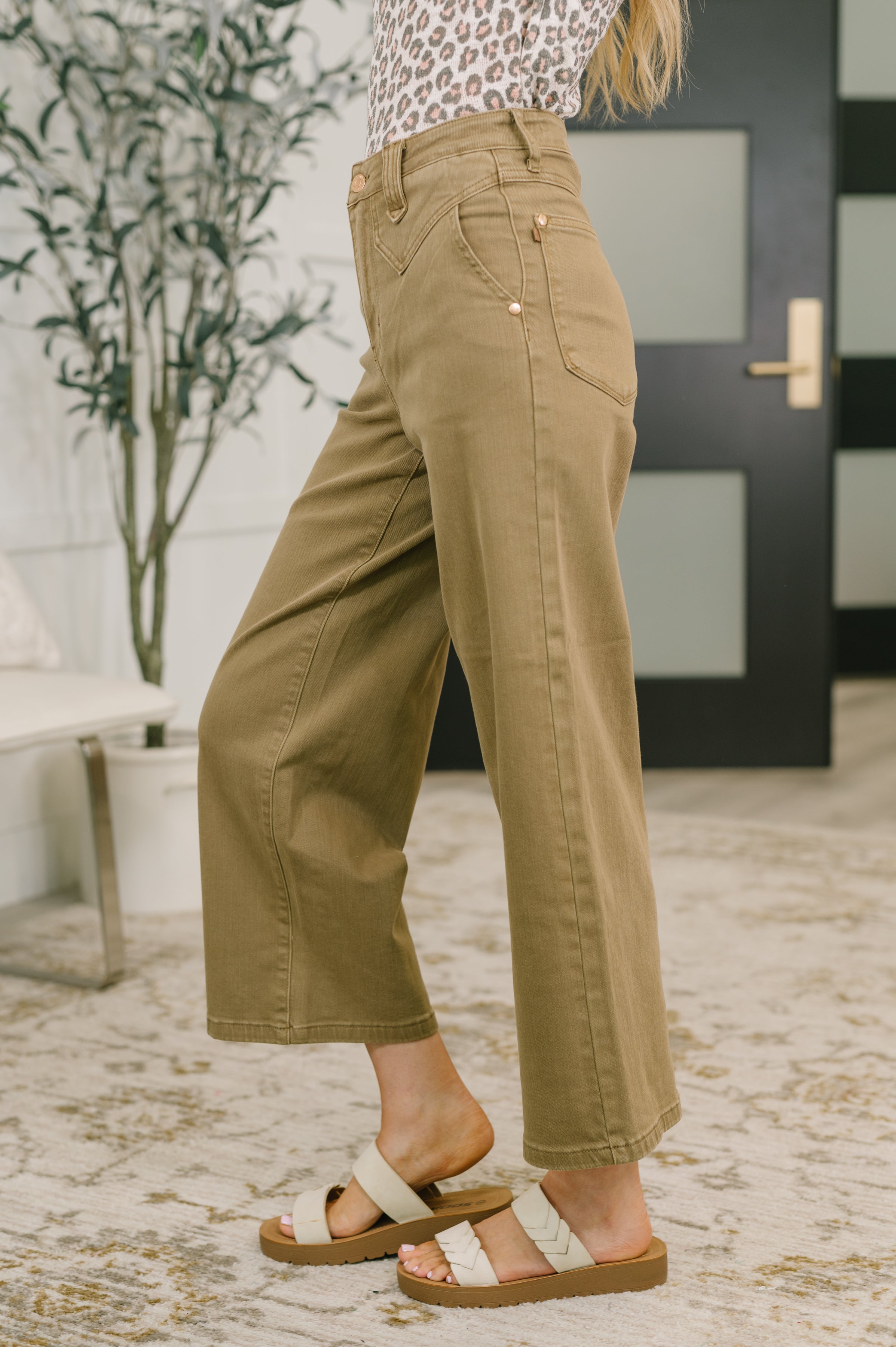 Side view of brown high-rise wide-leg ankle jeans with vintage front yoke paired with cream braided sandals indoors